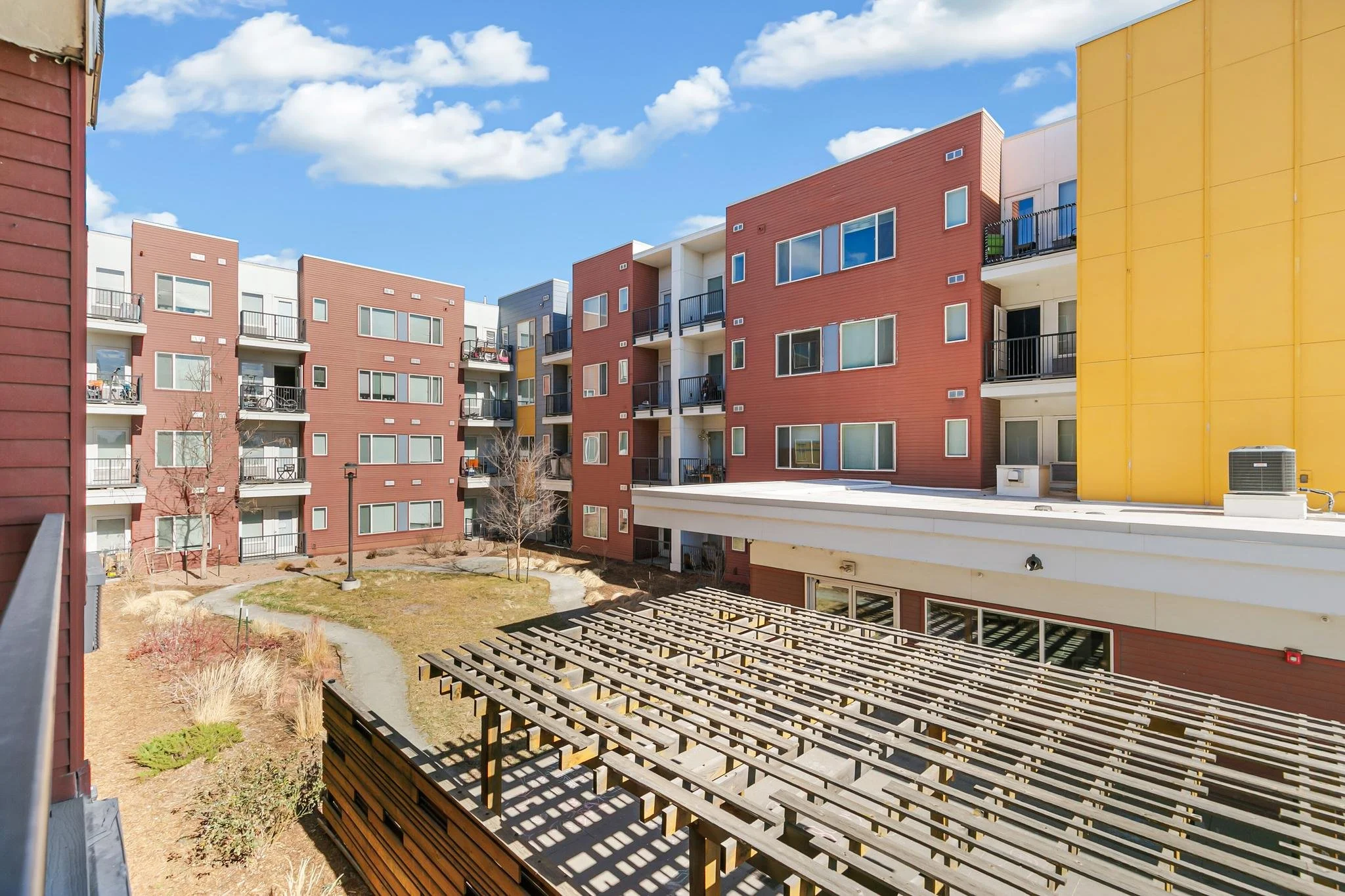 View of modern multi-story apartment buildings with colorful facades in red, white, gray, and yellow, featuring balconies, large windows, and a courtyard with a small pathway, trees, and outdoor furniture, under a partly cloudy sky.