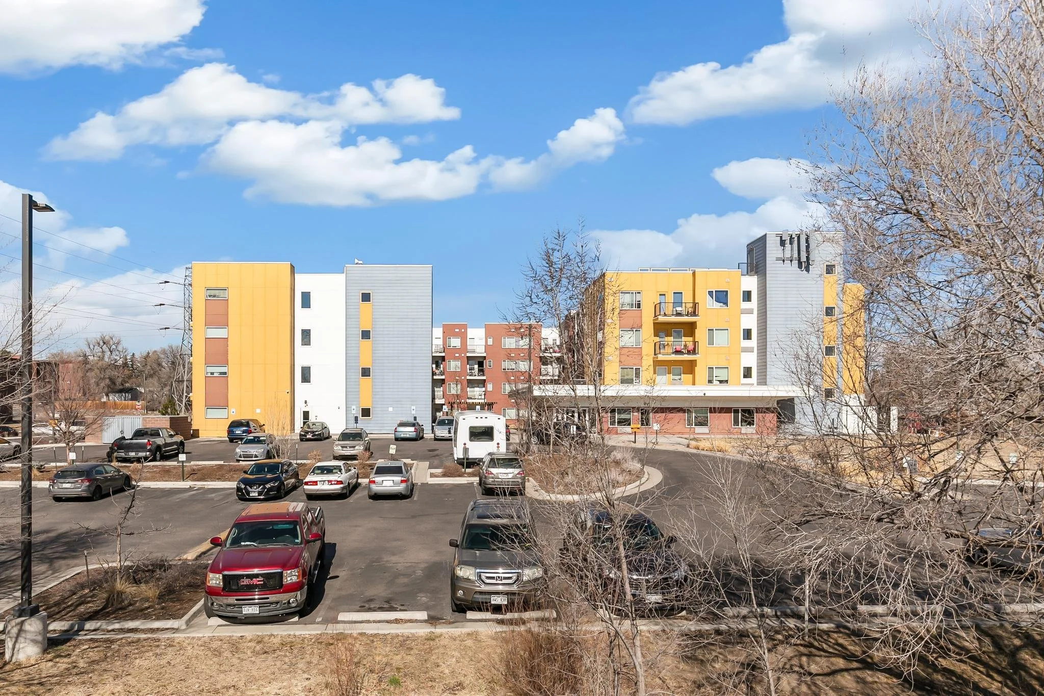 Colorful multi-story apartment buildings with a parking lot in front, cars parked, leafless trees, blue sky with clouds, and lamp posts.