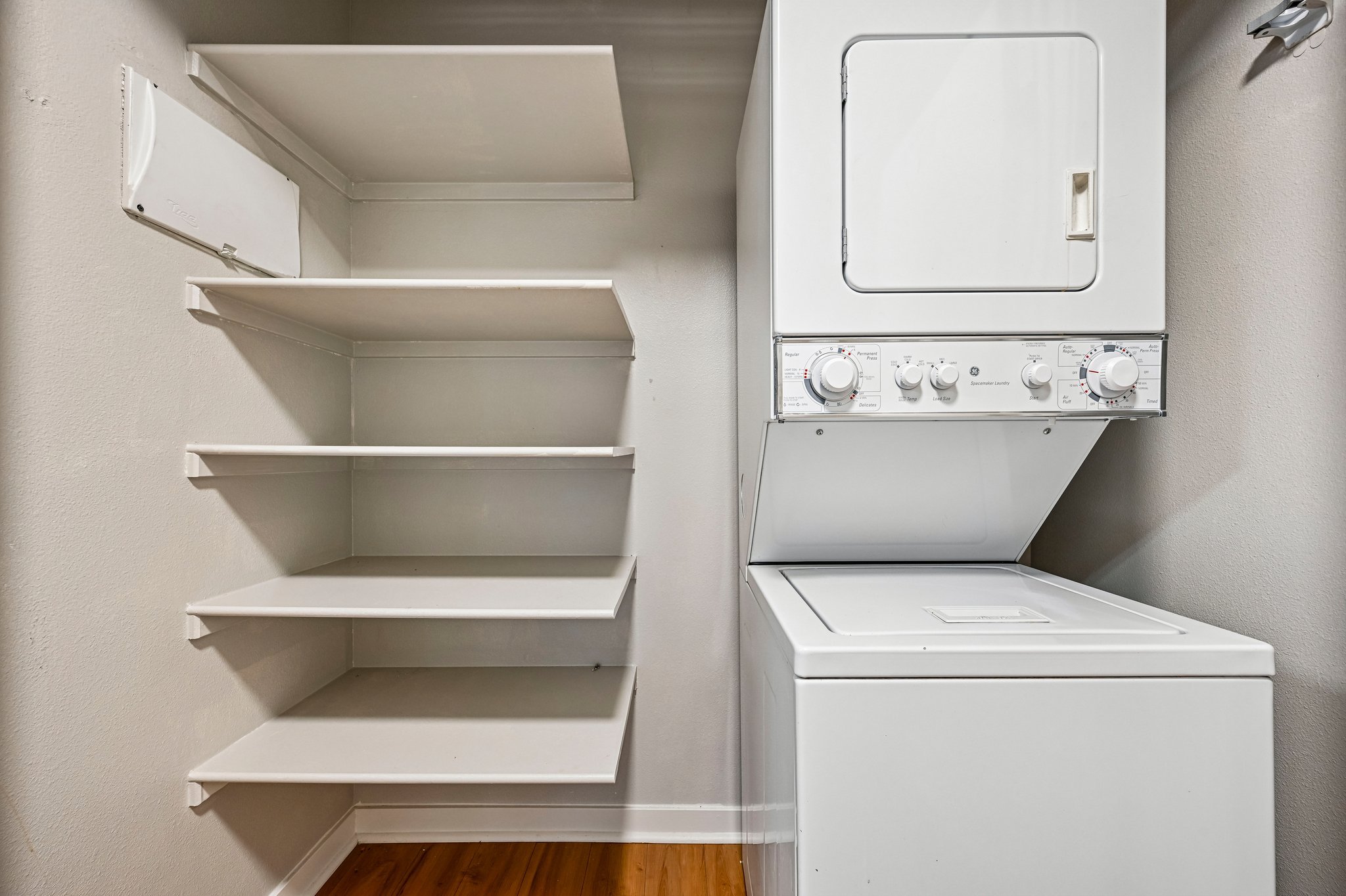 A laundry room with white walls and a wooden floor, featuring a white washing machine and dryer stacked on top of each other. There are four empty white shelves on the left wall.