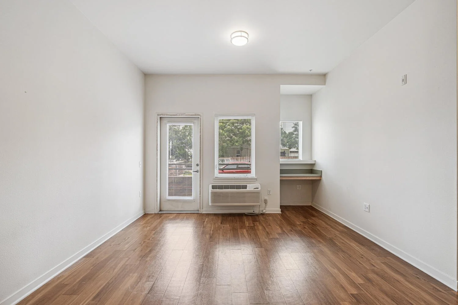 Empty room with hardwood floors, white walls, and a large window and door leading outside.