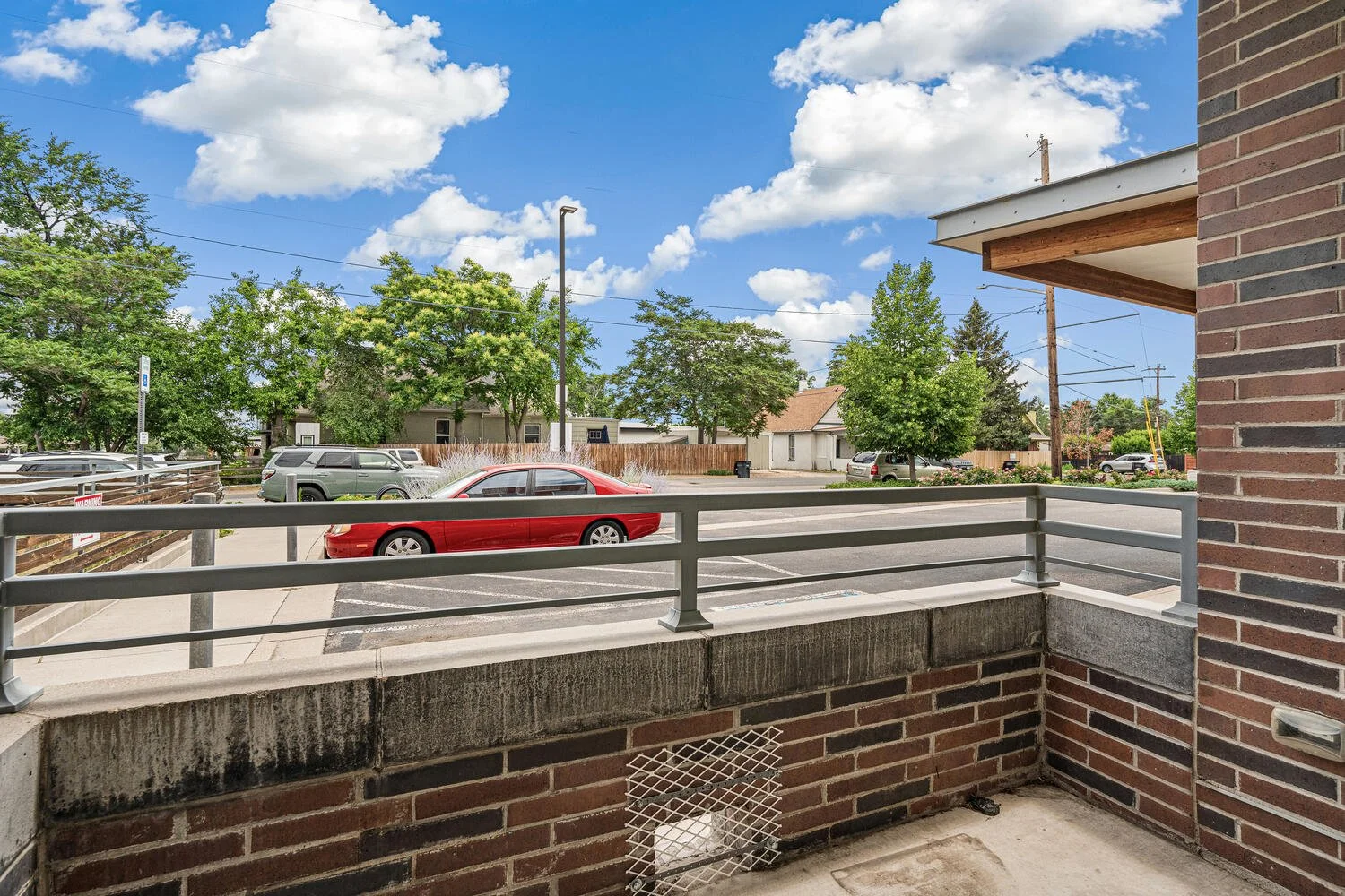 View from a brick balcony overlooking a parking lot with several cars, trees, and a blue sky with white clouds.