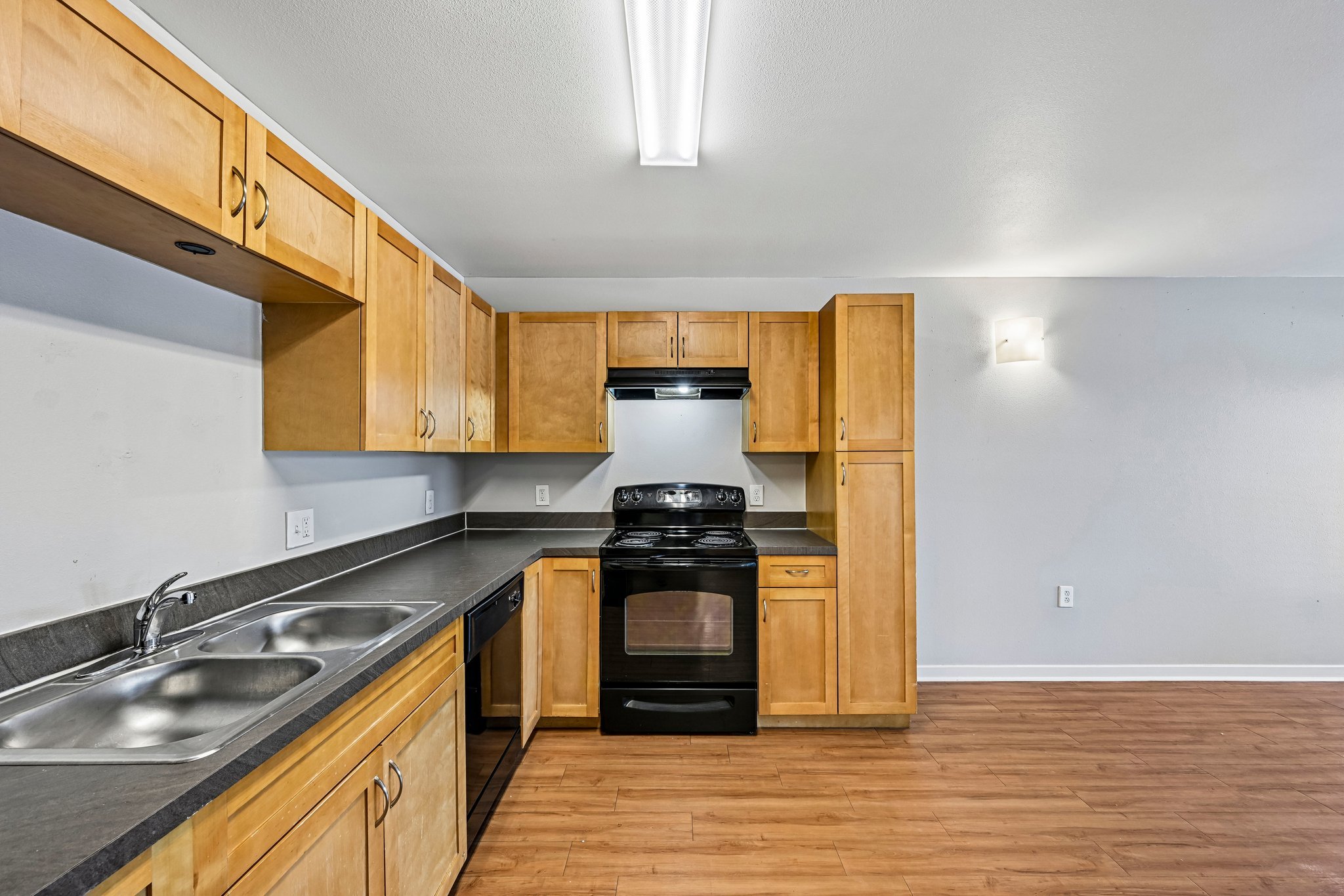 A kitchen with wooden cabinets, a black stove, and countertops with a double sink. The room has hardwood floors, a white wall, and ceiling lighting.