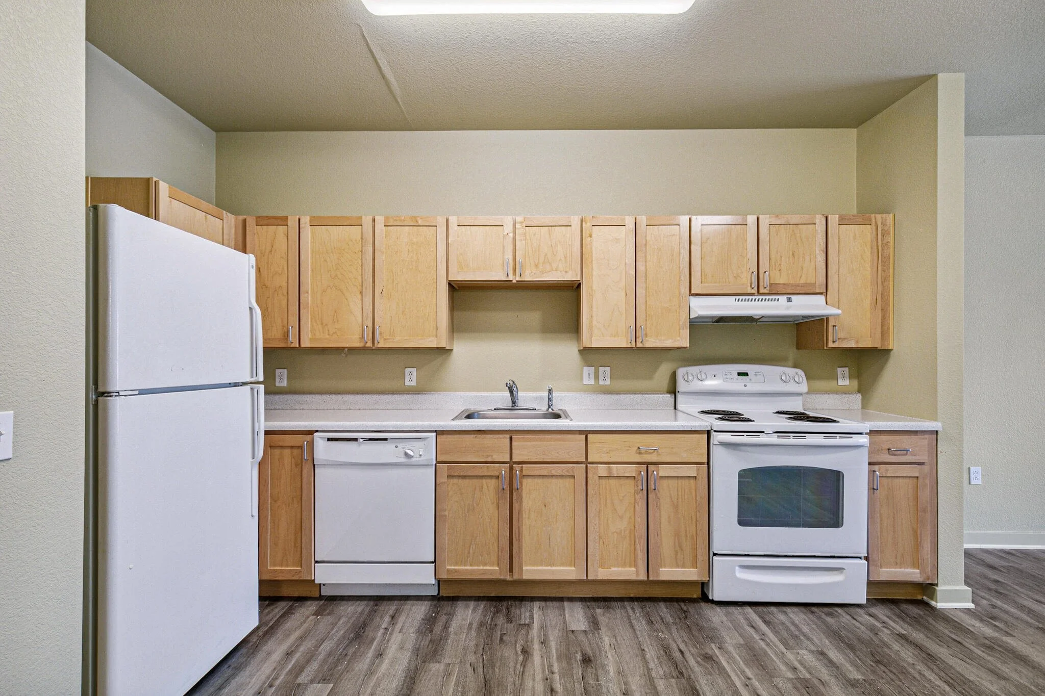 Kitchen with wooden cabinets, white appliances including a refrigerator, stove, and dishwasher, and a stainless steel sink. The floor has dark wood-like vinyl planks, and the walls are painted light beige.