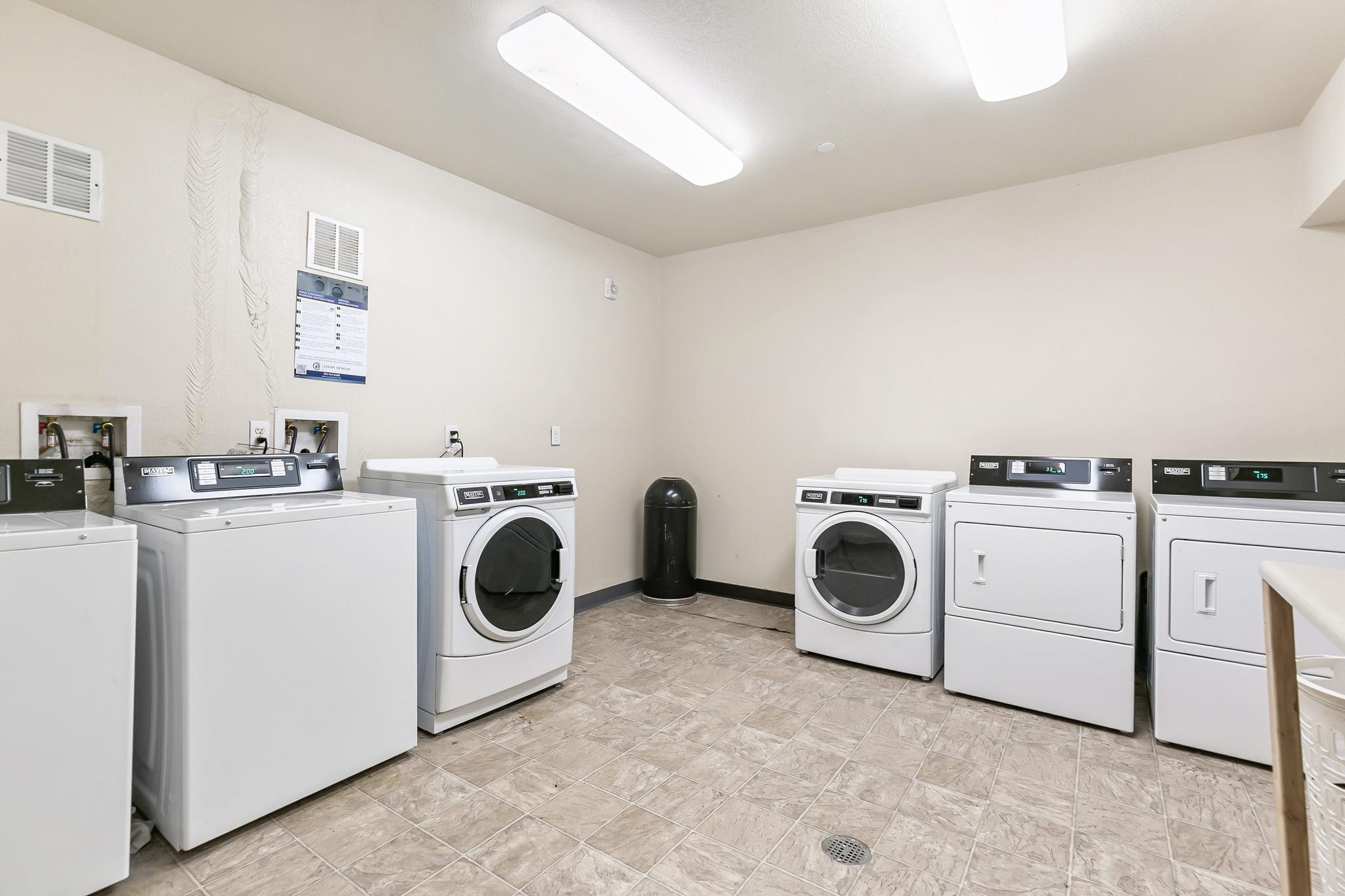 A laundry room with five white washing machines and one white dryer lined up against the wall, with vents and a sign on the wall, a black trash can, and a tiled floor.