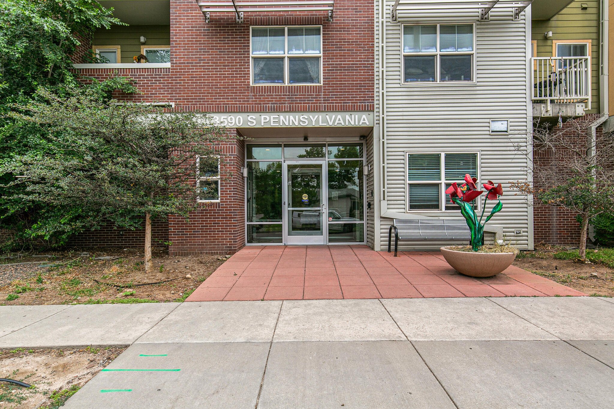 Exterior view of a multi-story residential building with a brick facade and a small outdoor patio area with red paving stones, trees, shrubs, and a colorful flower sculpture in a round planter.