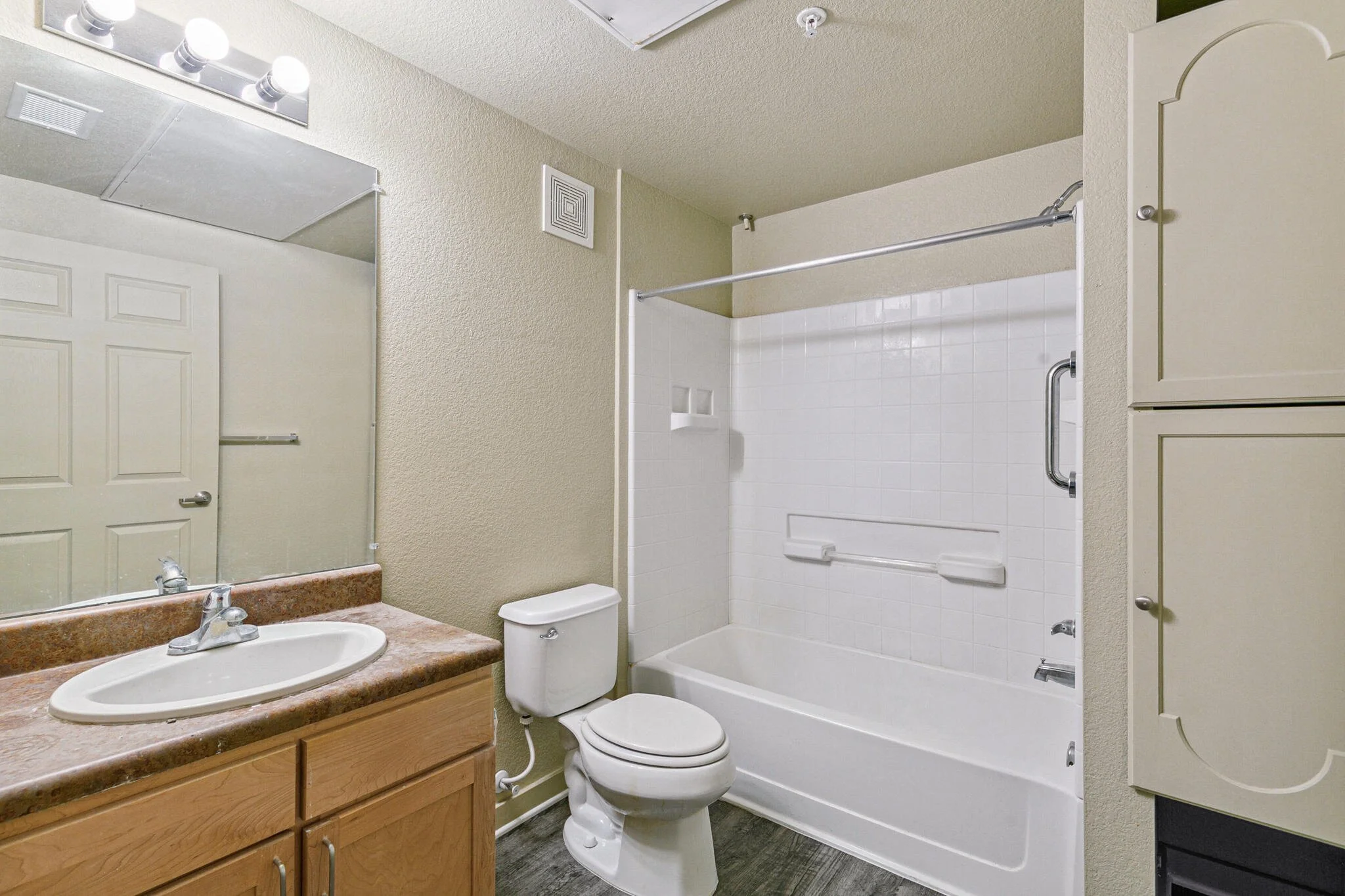 A small bathroom featuring a white bathtub with a showerhead and sliding shower doors, a white toilet, a wooden vanity with a sink and marble countertop, a mirror above the sink, a medicine cabinet, beige textured walls, a vent in the ceiling, and a ceiling light fixture.