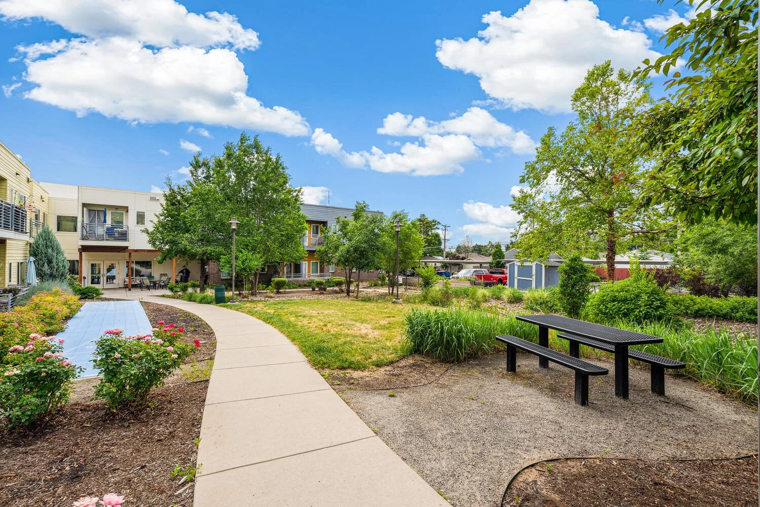 A well-maintained courtyard in a residential area with a concrete pathway, green trees, flowering plants, outdoor seating, and a clear blue sky with scattered clouds.