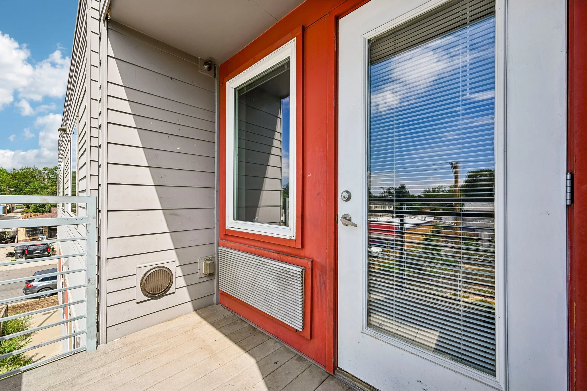 Balcony with a white door and large window with blinds, beige siding, and a red trim around the window and door. Outside, there are parked cars and trees under a partly cloudy sky.