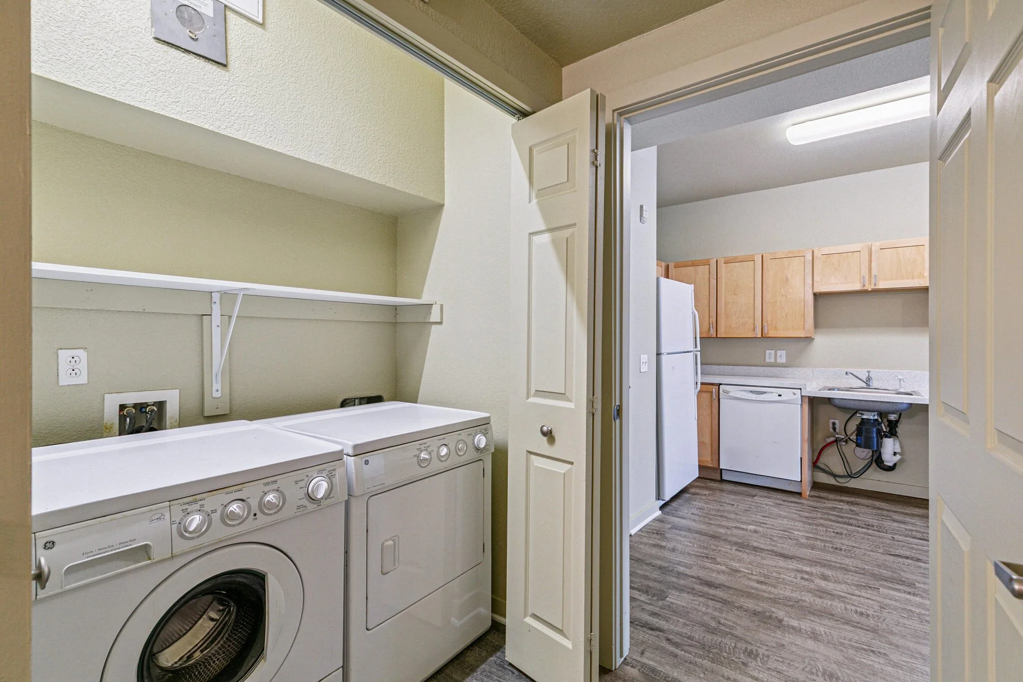 View into a laundry room with two washers, a clothes dryer, and a small laundry sink in the background. The room also has a small countertop, cabinets, and a wall with a power outlet and water hookups.
