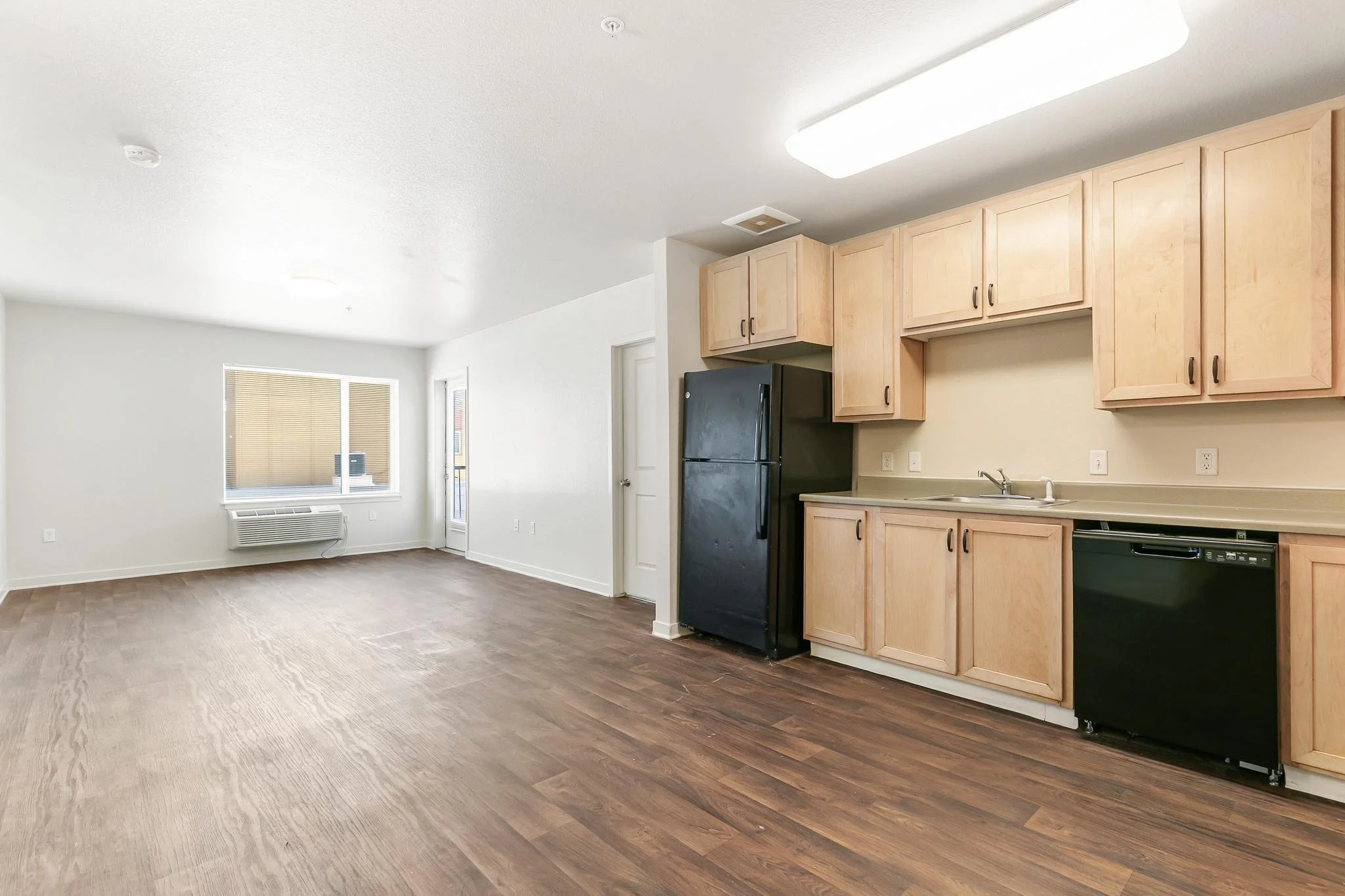 Interior of an unfurnished apartment with an open-concept living room and kitchen, featuring wooden flooring, white walls, a window with blinds, and light wood kitchen cabinets.