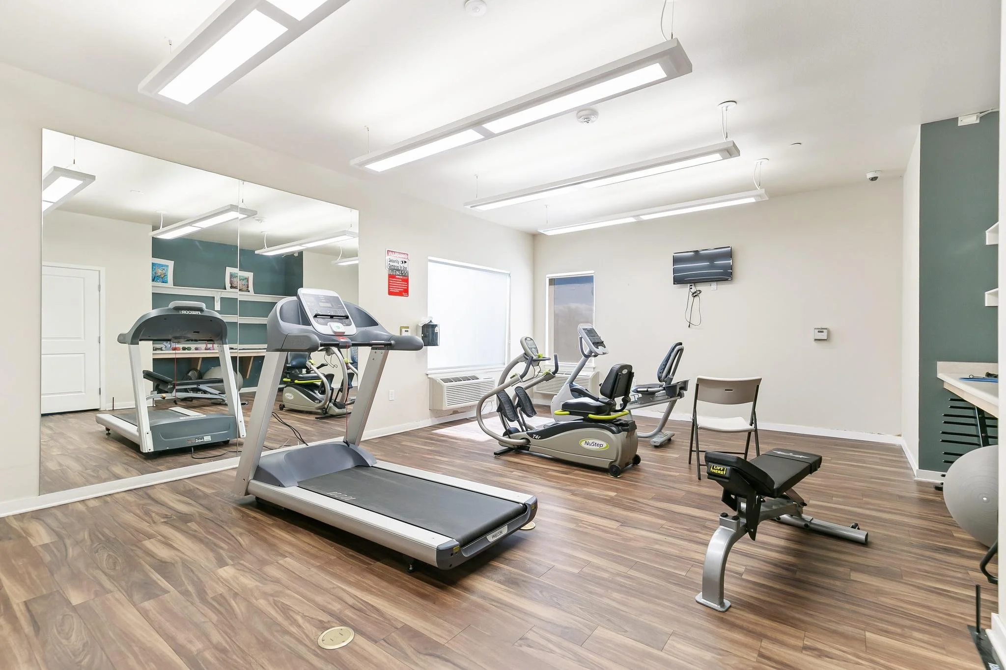 A fitness room with treadmills, exercise bikes, and other gym equipment on wood-style flooring, with large mirrors and a mounted flat-screen TV on the wall.