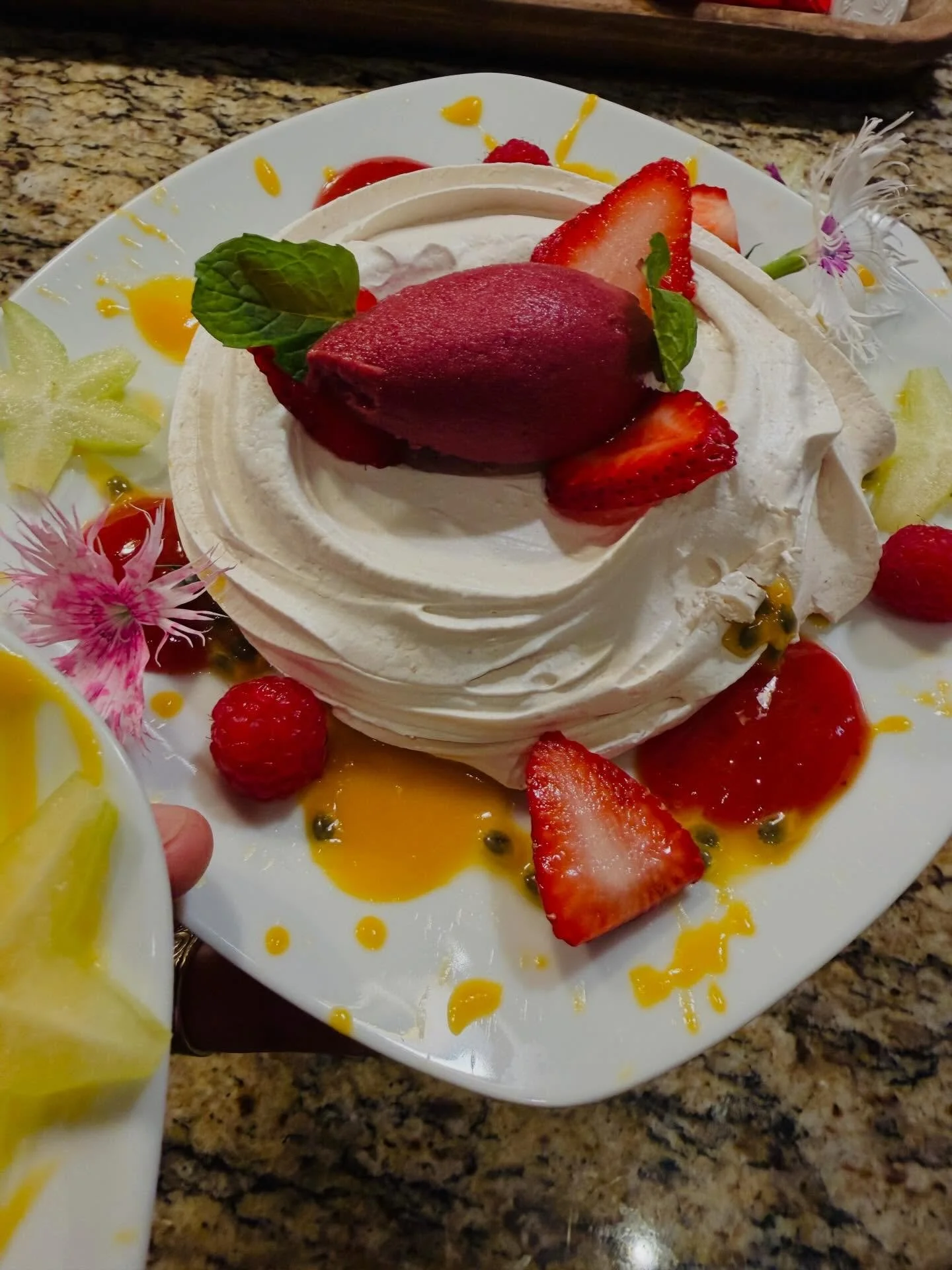 An elegant island-inspired pavlova with a crisp, snowy meringue shell and a soft, marshmallowy center, topped with silky whipped cream and a quenelle of vibrant berry sorbet. Finished with fresh strawberries, raspberries, passion fruit, mango coulis,