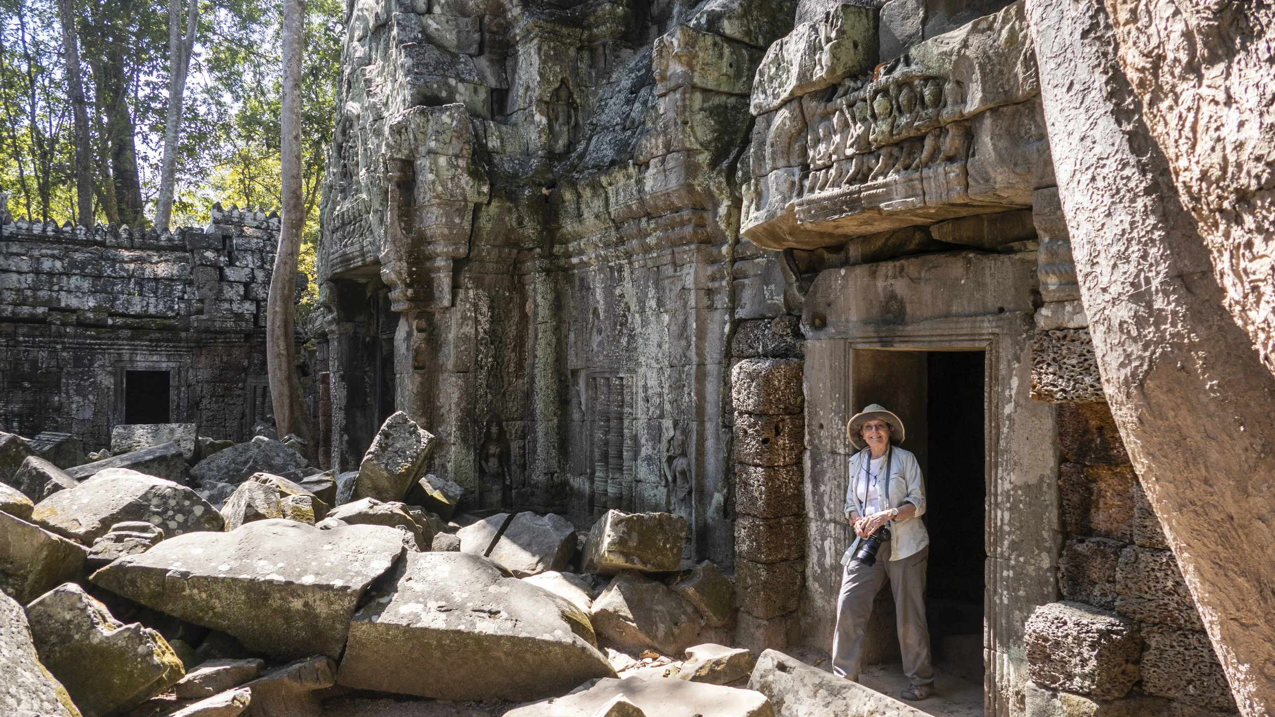 A woman standing at the entrance of an ancient Cambodian stone temple surrounded by fallen rocks, with intricate carvings on the stone walls, and trees in the background.