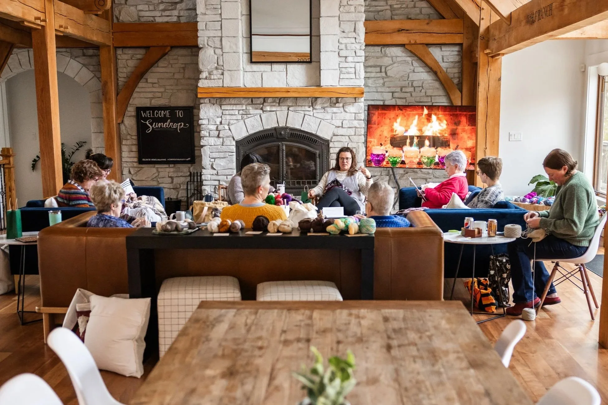 A group of people gathered in a cozy living room, sitting on various chairs and sofas in front of a fireplace, with yarn and knitting supplies on tables and shelves, participating in a knitting or craft workshop.