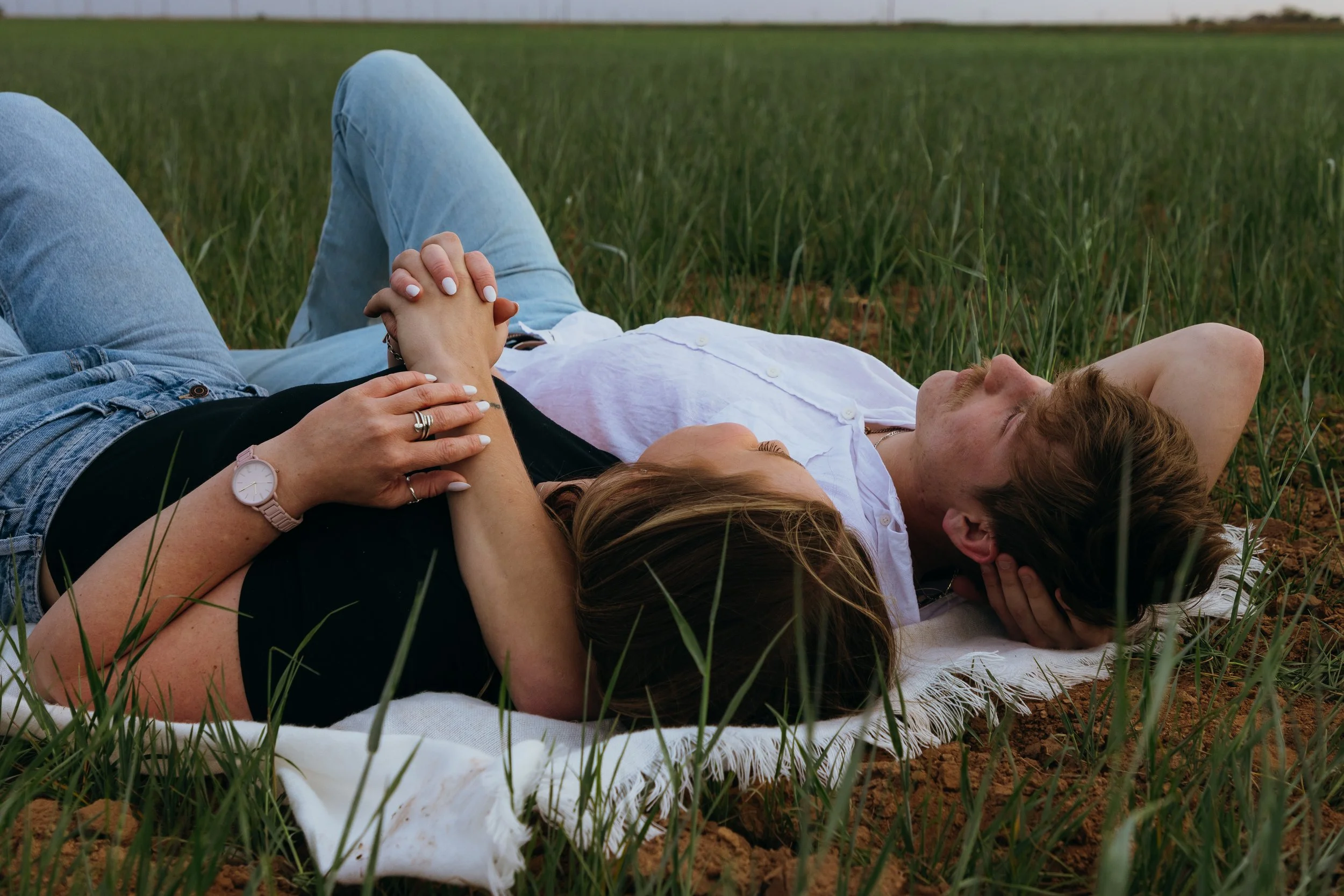 Couple lying on a blanket in a grassy field holding hands, candid outdoor moment, portrait photography