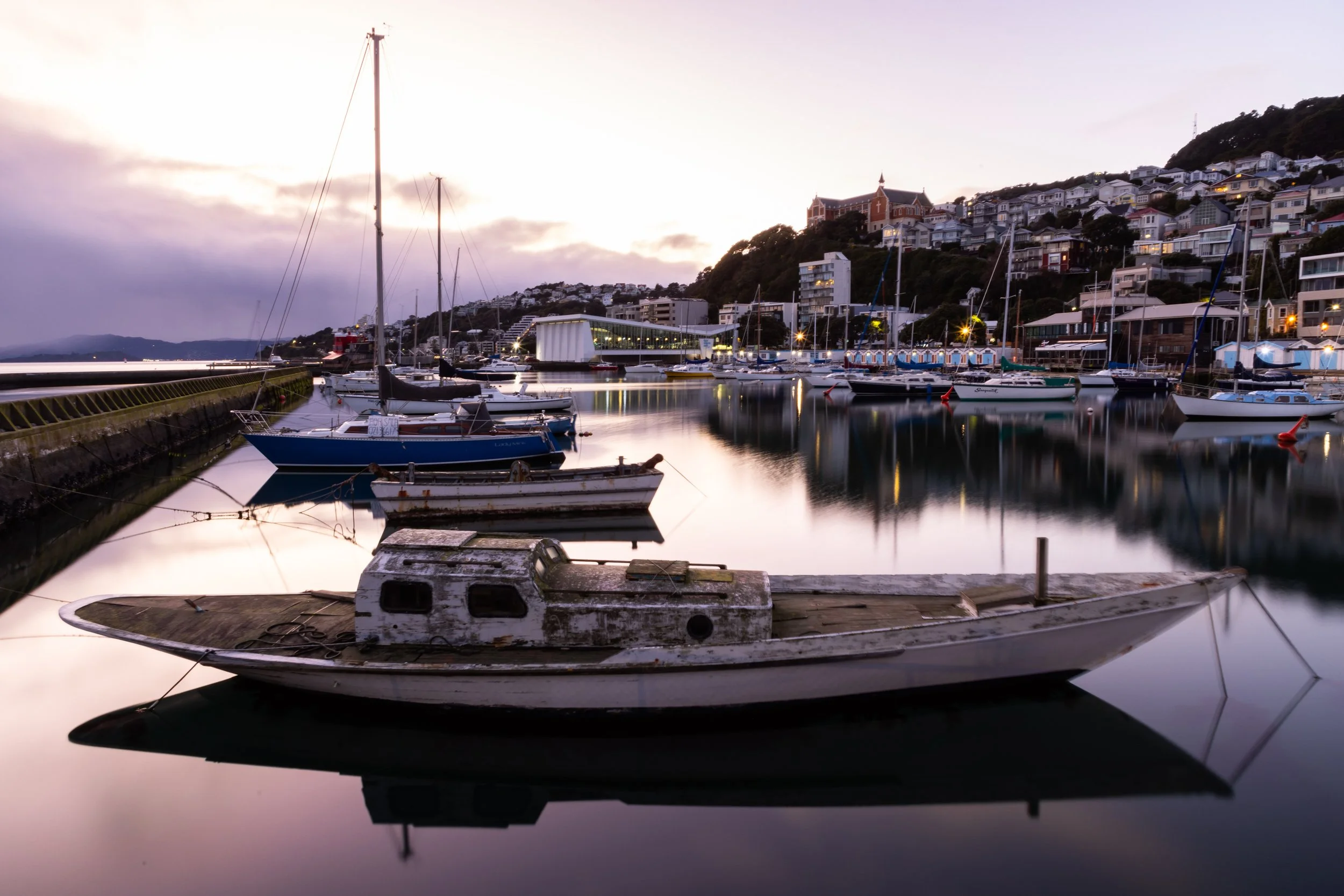 Coastal marina at sunrise with sailboats and hillside town, calm harbor reflections