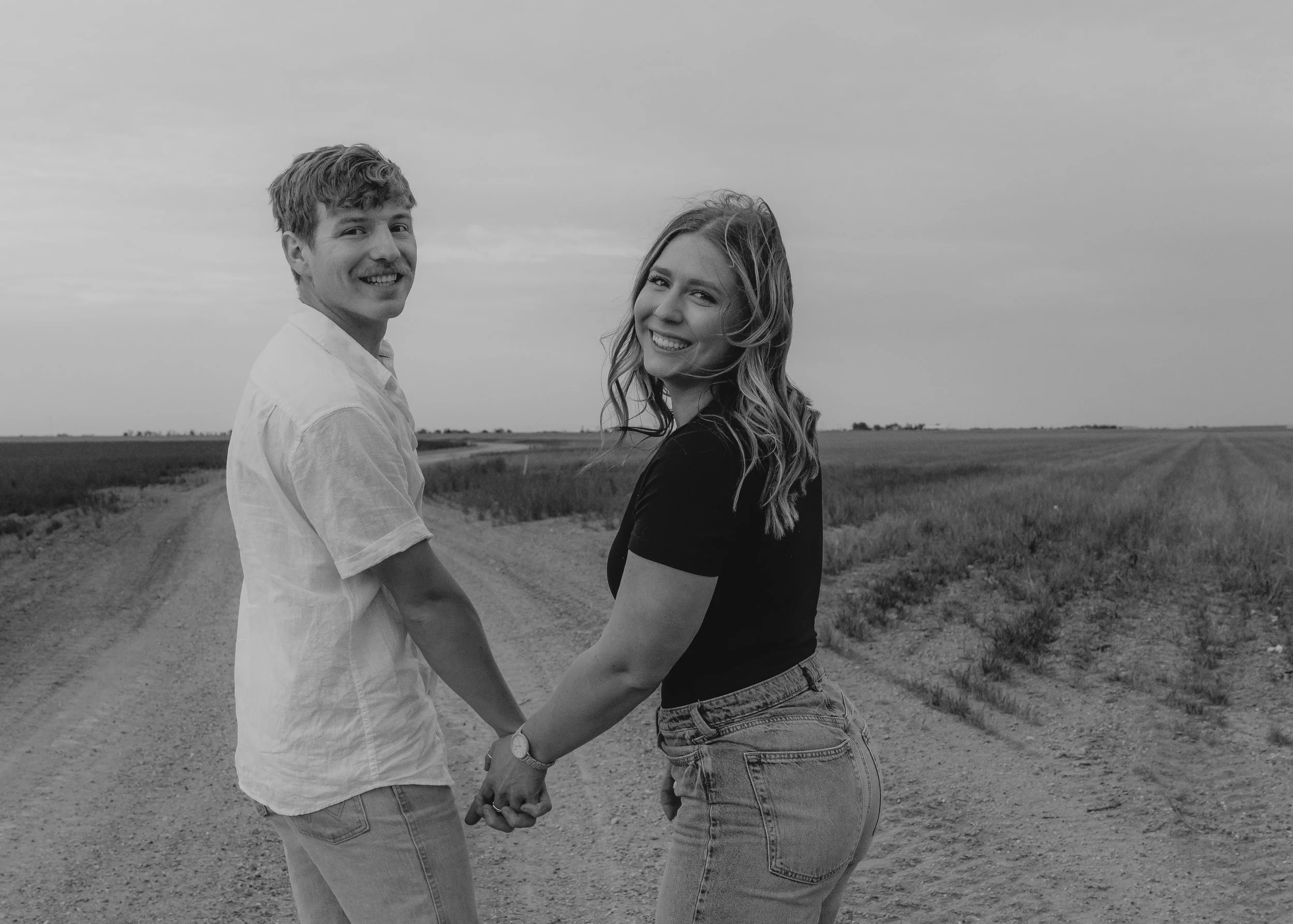 A smiling couple holding hands and walking down a dirt road in a rural area, with open fields