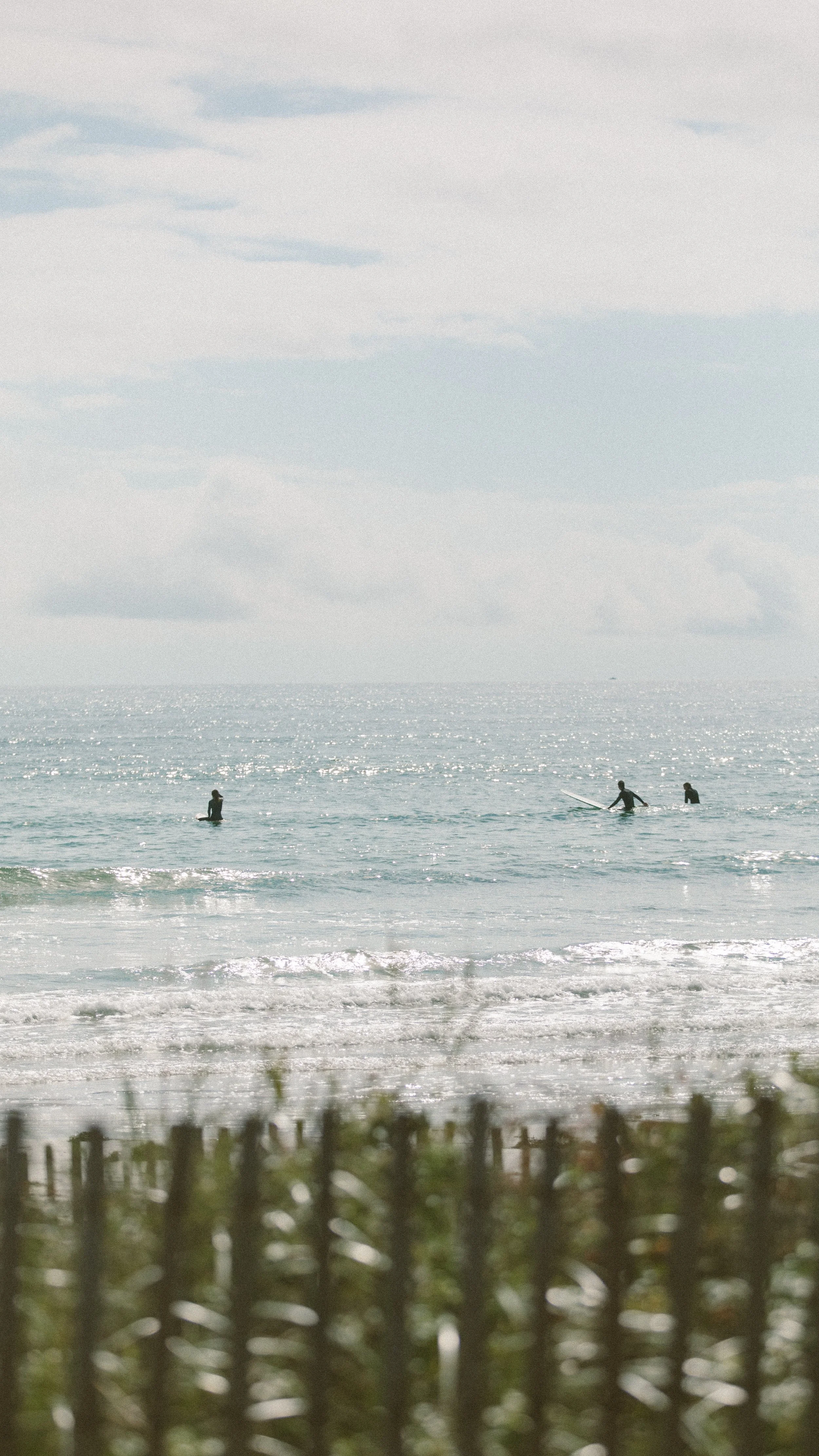Three people in the ocean near the shore, one standing and two on surfboards, with a cloudy sky above and a wooden fence in the foreground.