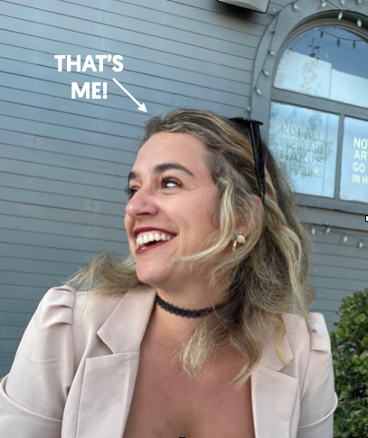 A woman with blonde hair smiling and wearing a beige blazer, choker necklace, and sunglasses on her head. The background is a gray building with a round window. An arrow and text label point to her saying, "THAT'S ME!"