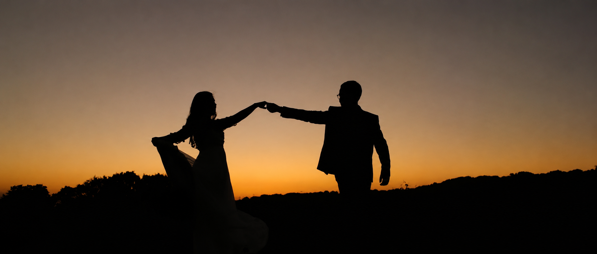 Silhouettes de deux personnes, un homme et une femme, dansant ou se tenant la main lors du coucher du soleil dans un paysage naturel.