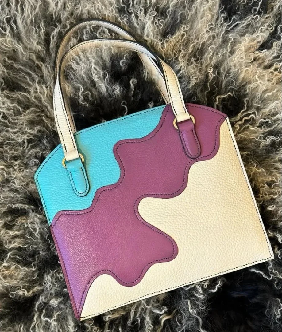 A colorful handbag with blue, purple, and cream sections, featuring an abstract design, placed on curly black and white fur.