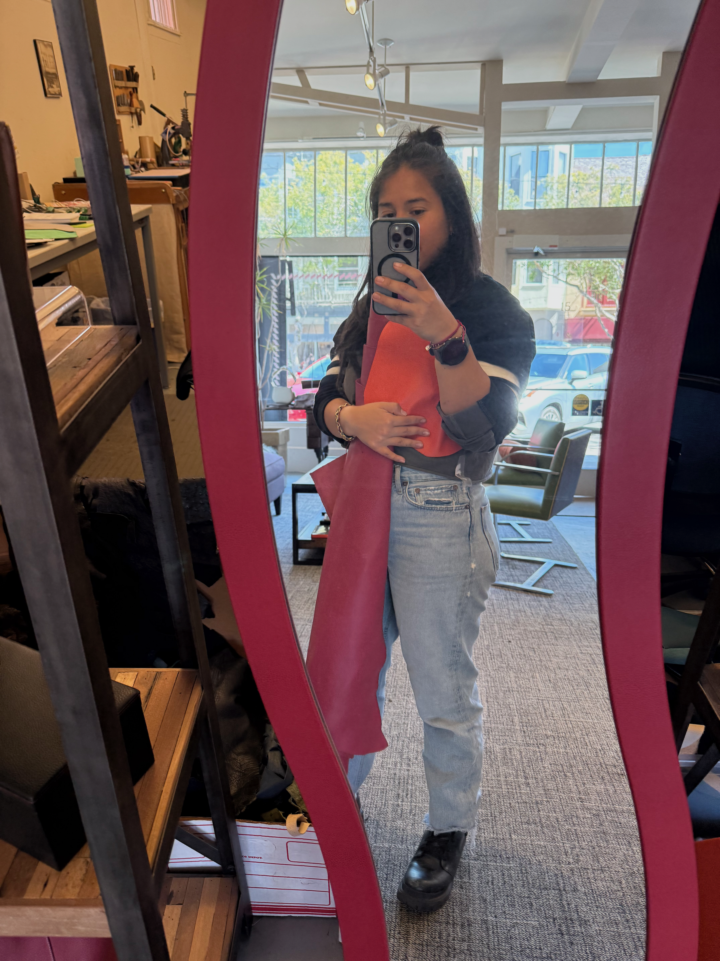 A young woman taking a mirror selfie inside a store. She is wearing a dark jacket, orange top, light blue jeans, and black boots. The mirror has a pink border, and the store has various furniture and objects in the background with large windows showing trees outside.