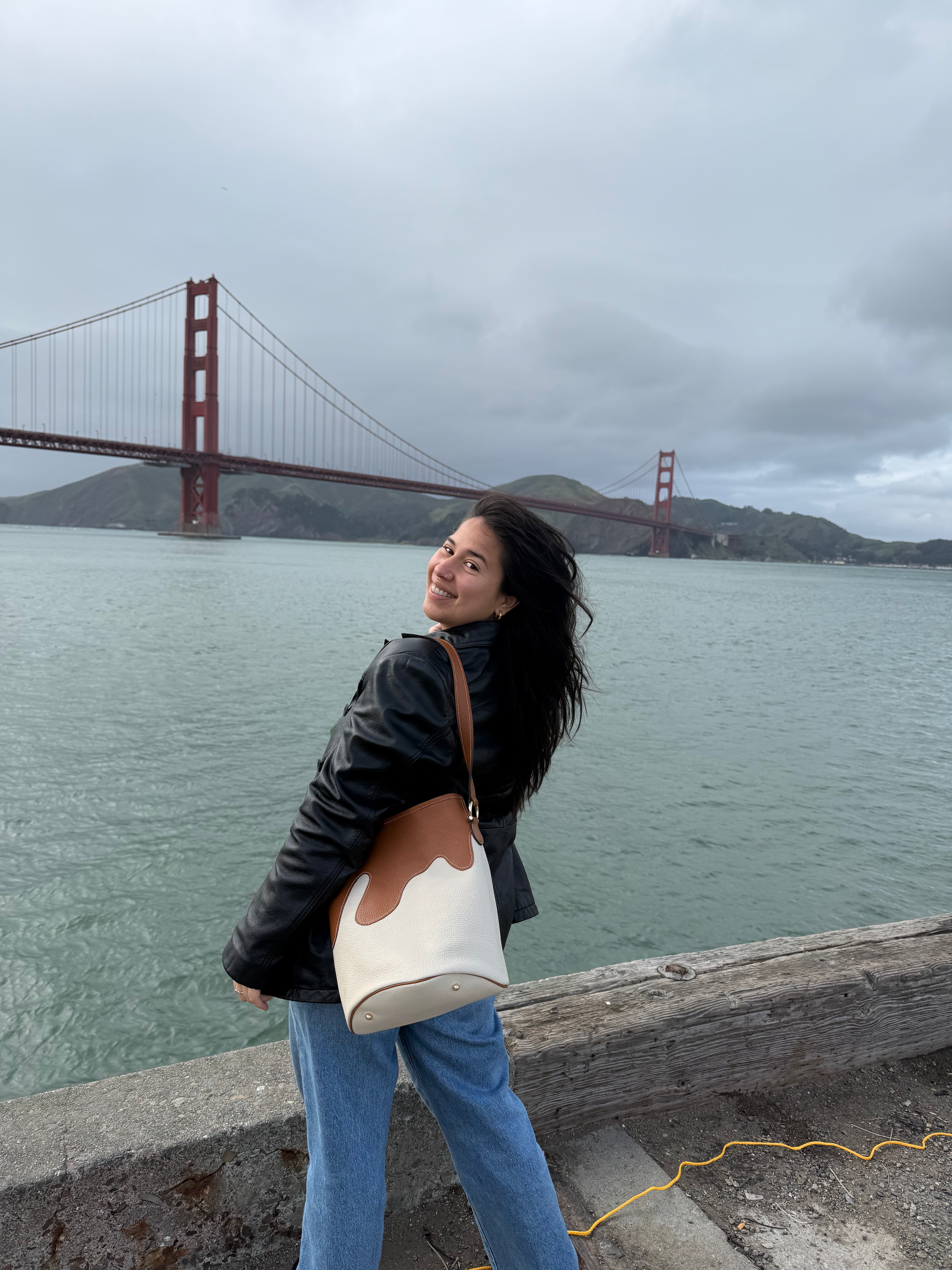 A woman with long dark hair smiling at the camera stands near a body of water with the Golden Gate Bridge in the background. She is wearing a black leather jacket and blue jeans, and carrying a large beige and brown bag.