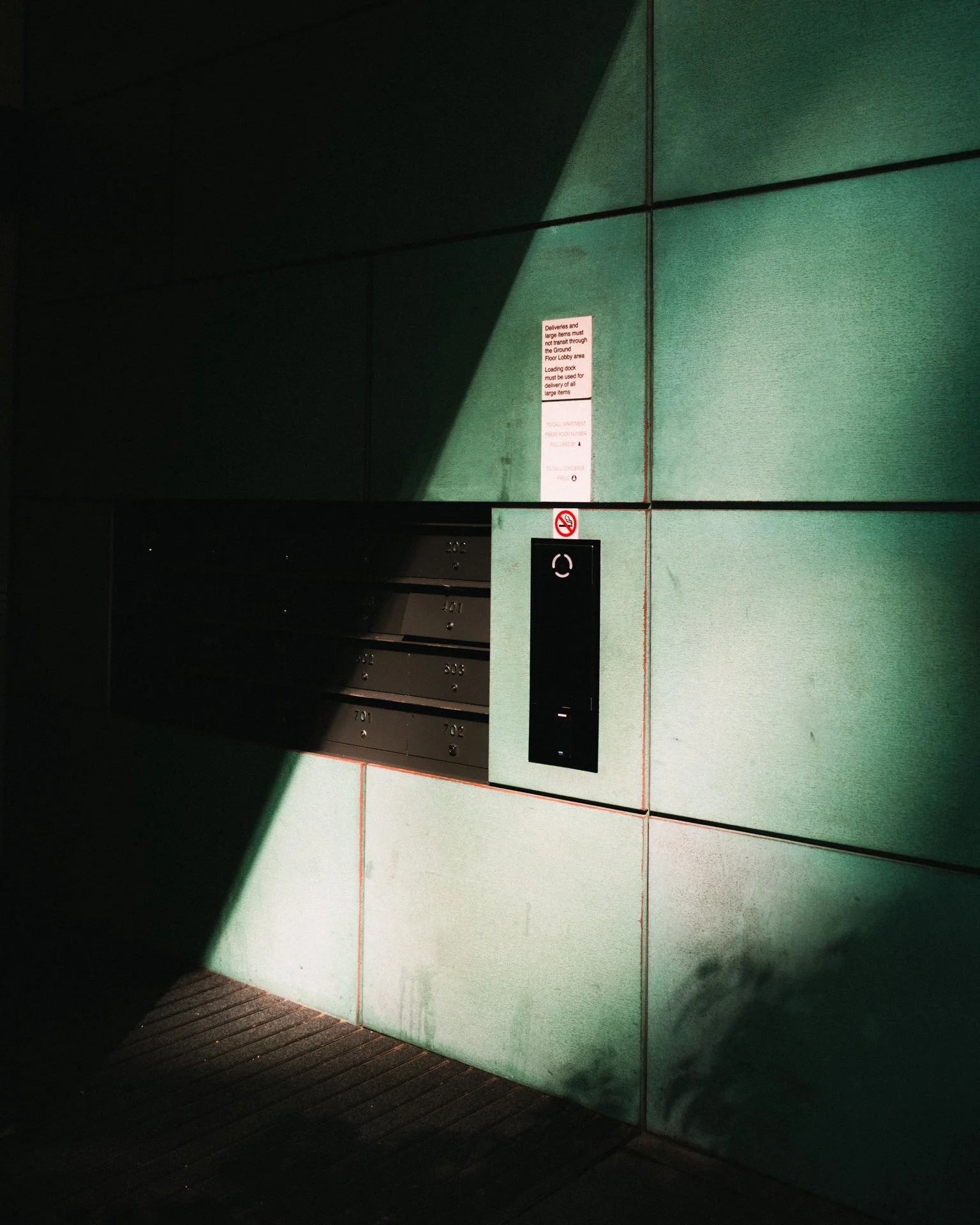 Green exterior wall with a row of mailboxes and an elevator panel, with shadows cast across the wall.