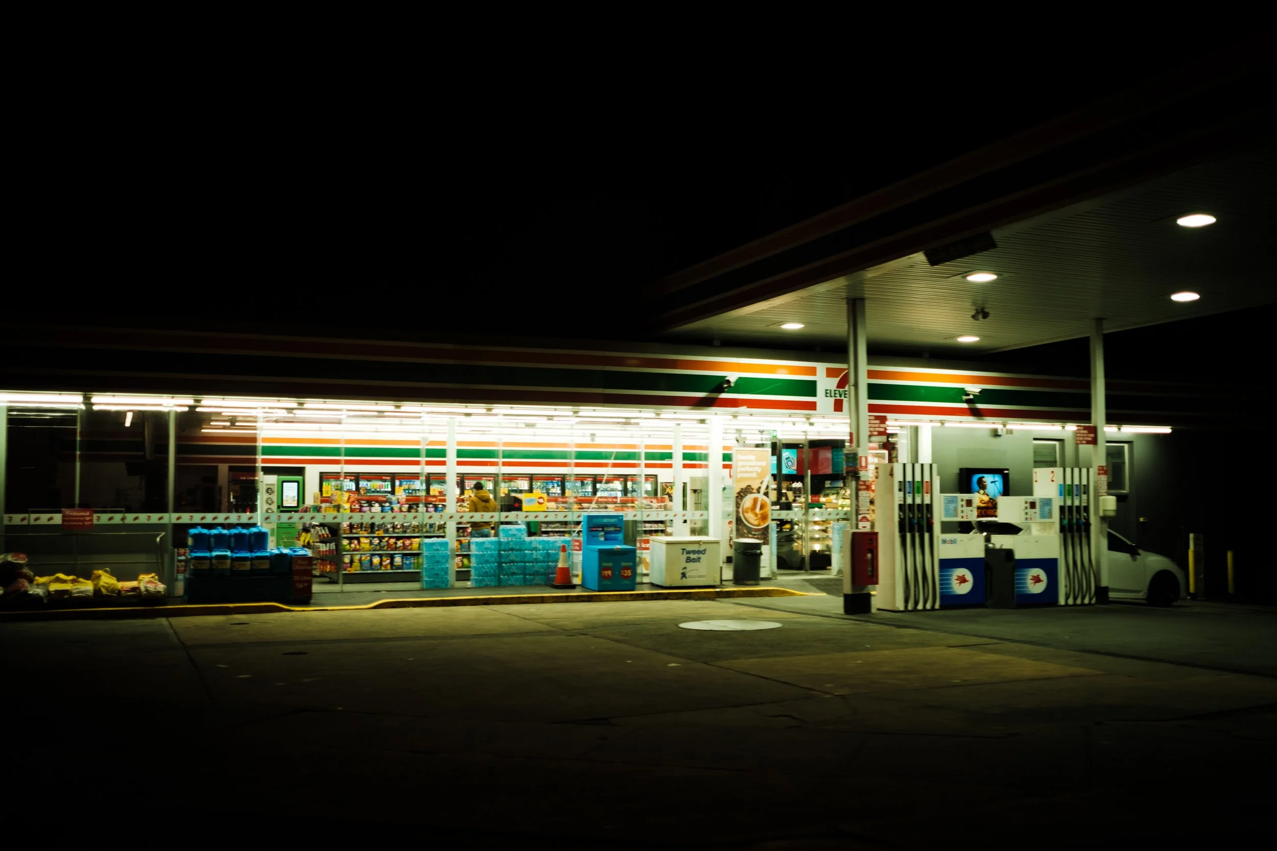 Nighttime scene of a 7-Eleven convenience store with illuminated signage, gas pumps, and a car parked to the right.
