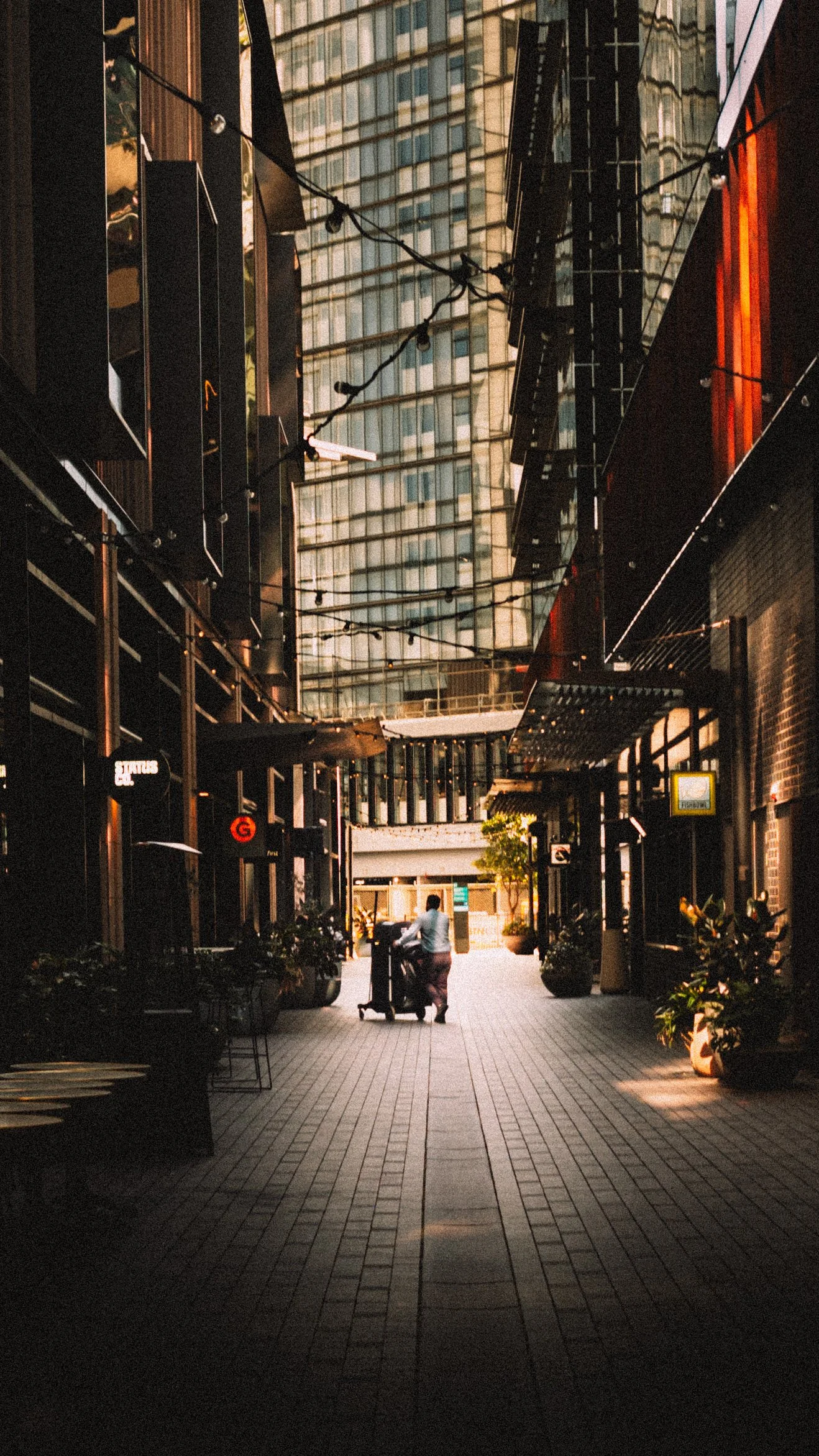 Urban alleyway with modern buildings, potted plants, and a person pushing a cart in the center, illuminated by warm lighting.