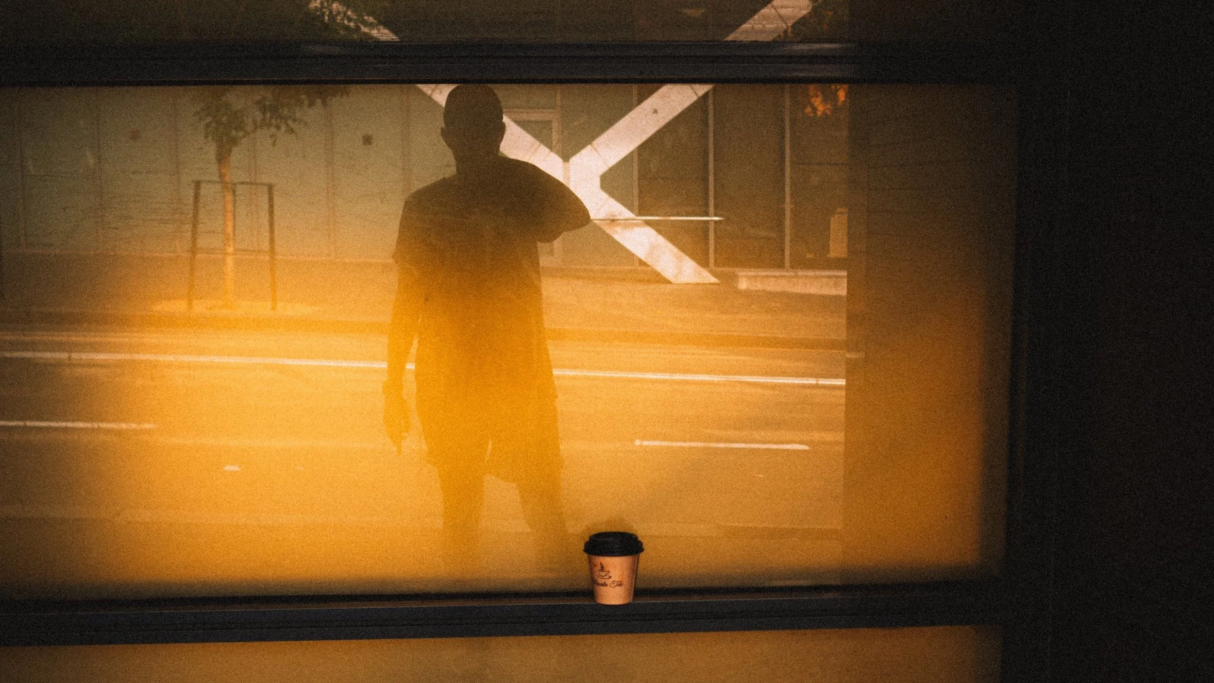 Silhouette of a man standing outside a window, with a coffee cup placed on the window sill. The background shows a fence and some trees.