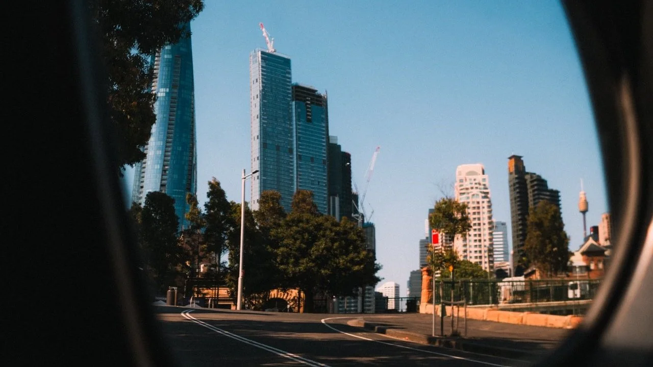 City skyline viewed through a car window, with tall buildings and construction cranes visible, during daytime.