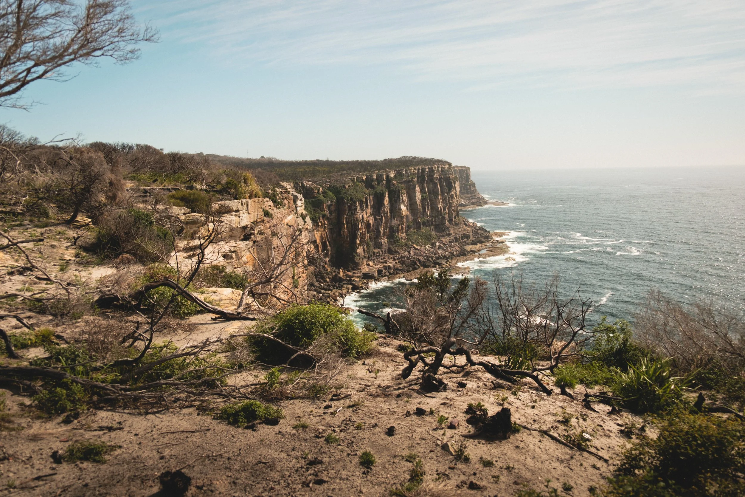 Cliffside view of a rugged coastline with steep, rocky cliffs and sparse, dry vegetation. The ocean waves crash against the rocks below, and a clear sky is visible above.