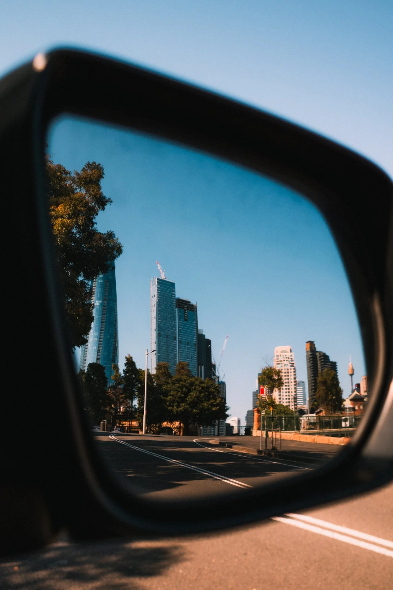 City skyline with tall buildings seen through a car's side mirror on an empty road with trees, under a clear blue sky.