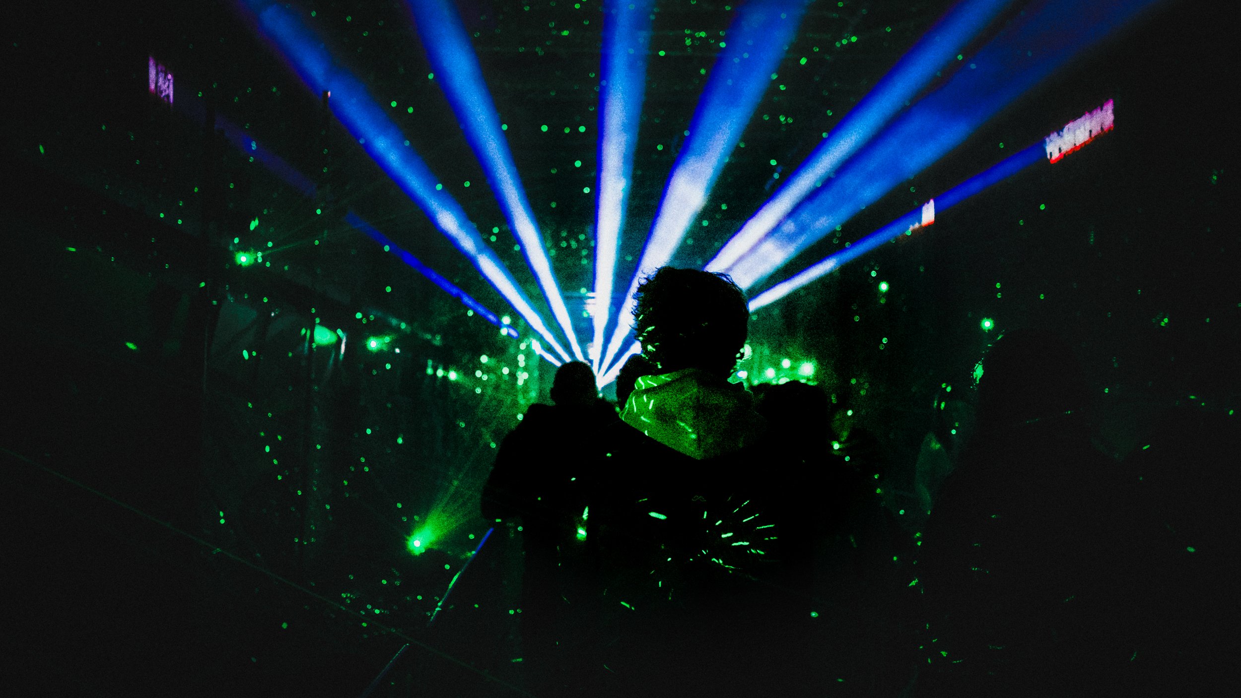 People are dancing in a dark club with green laser lights and blue spotlights creating a vibrant atmosphere.
