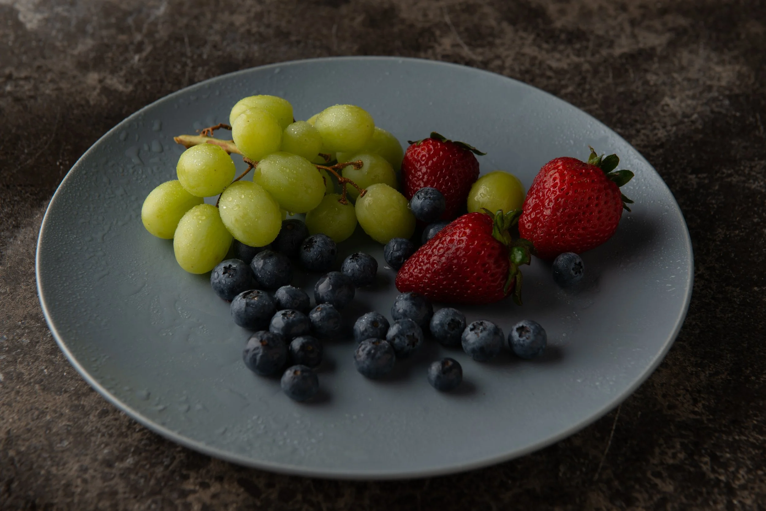A blue plate with green grapes, strawberries, and blueberries on a dark surface.