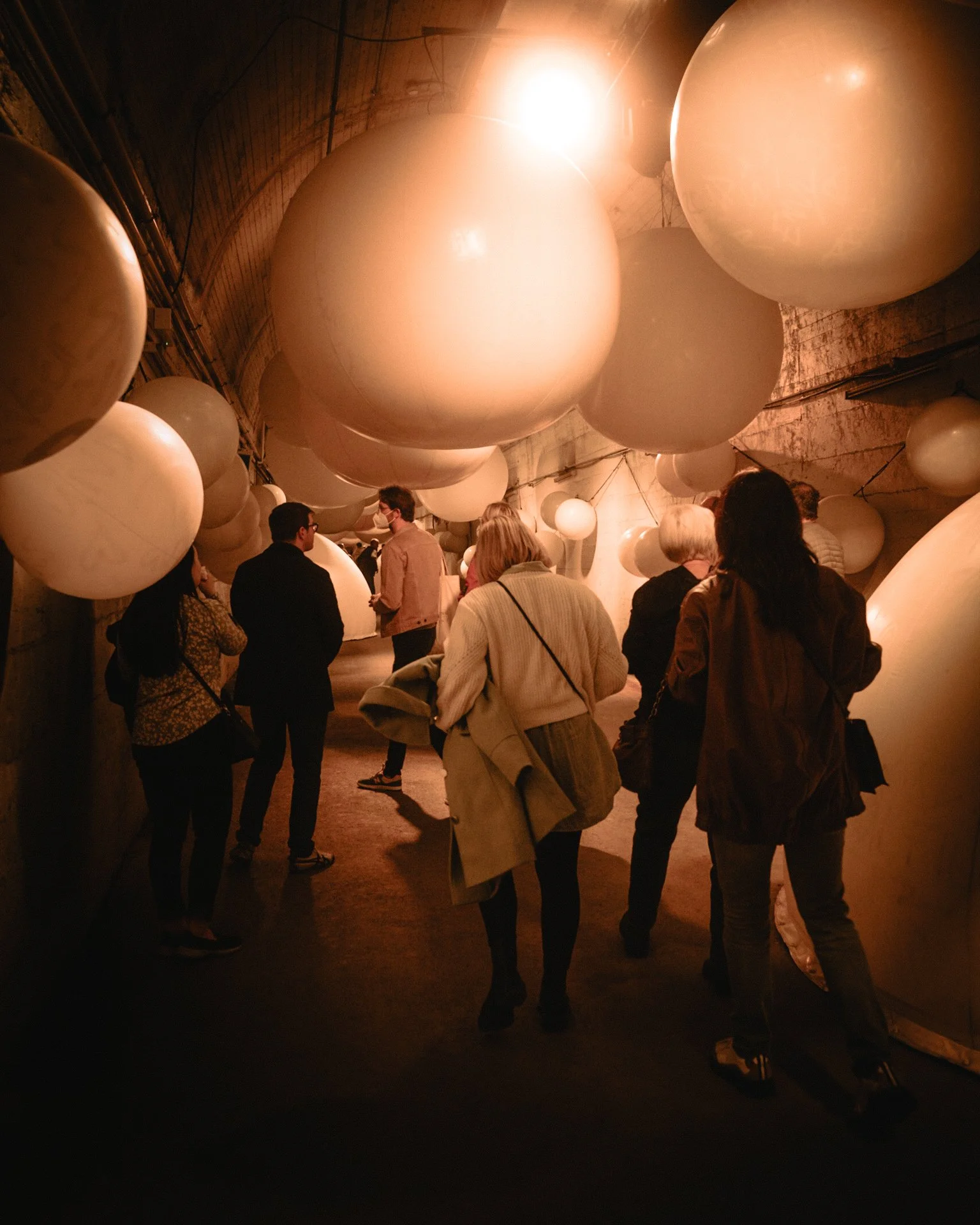 Group of people touring an art installation with large hanging white spheres in an underground tunnel.
