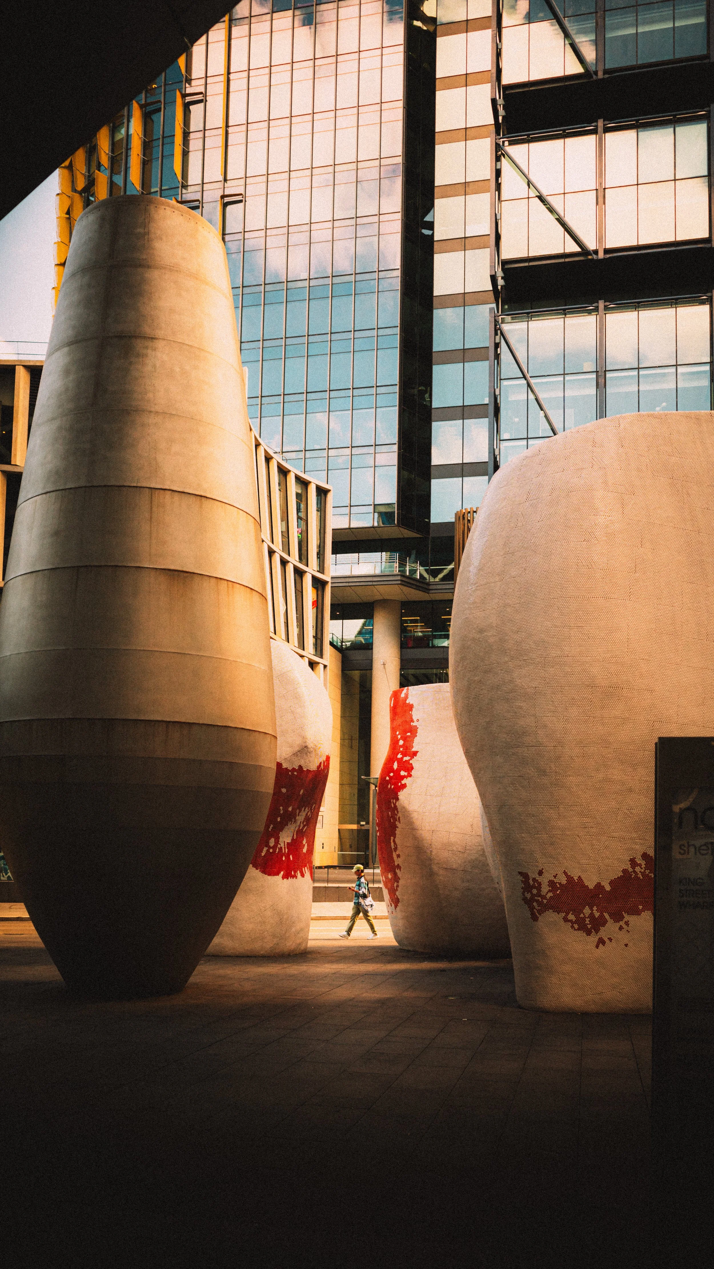 Large modern sculptures with abstract, rounded shapes and red patterns, located in an urban plaza with a glass building in the background, with a person walking in the distance.