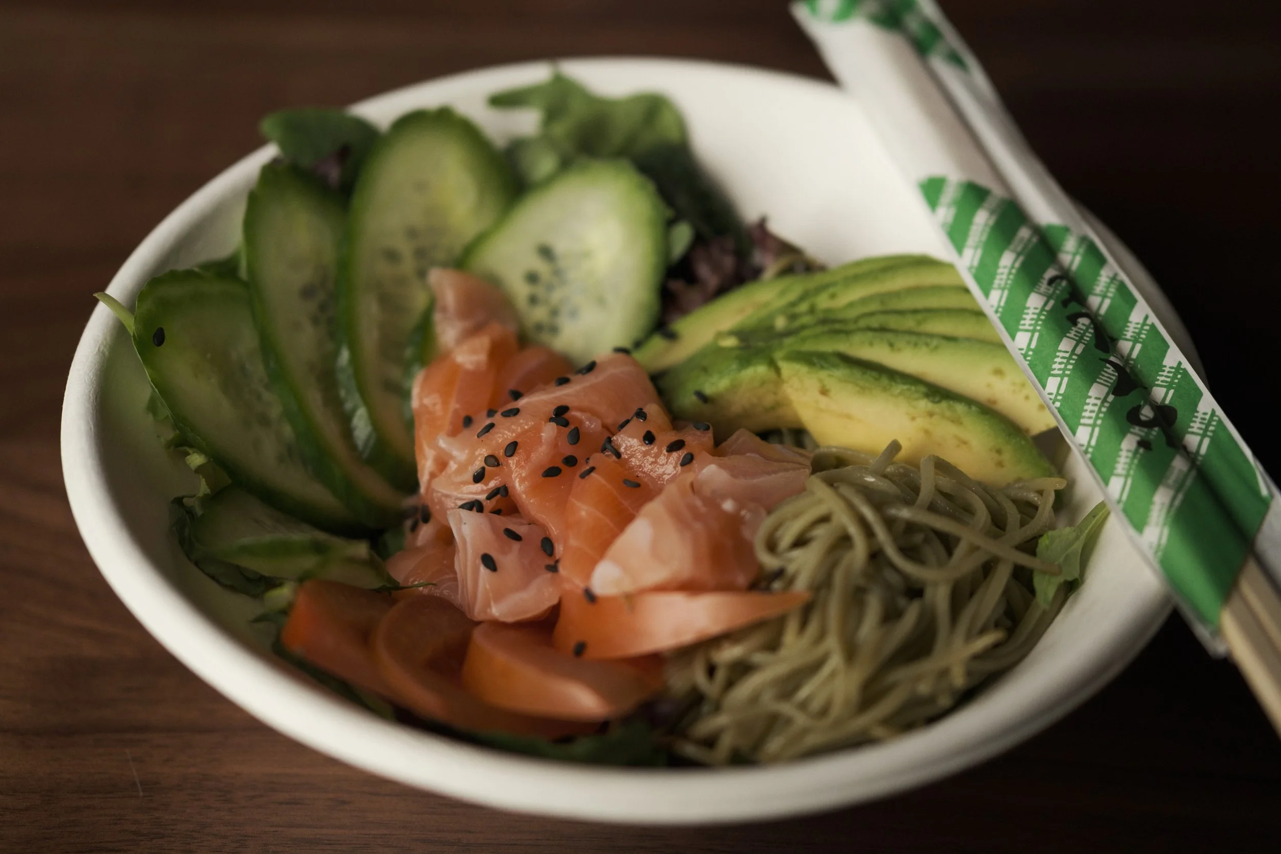A takeout bowl with sliced cucumbers, avocado slices, raw salmon topped with black sesame seeds, and noodles, with chopsticks resting on the side.