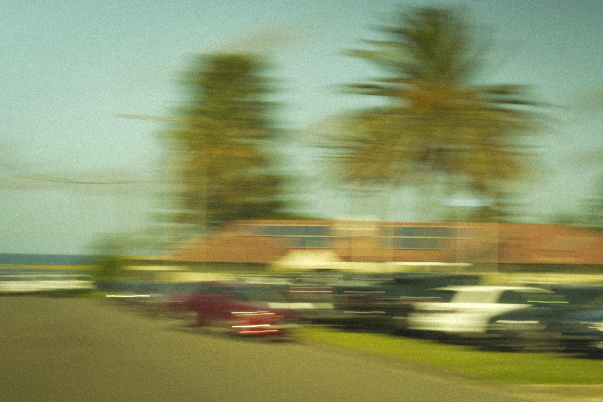 Blurred image of trees and parked cars along a street with a building and a clear sky in the background.