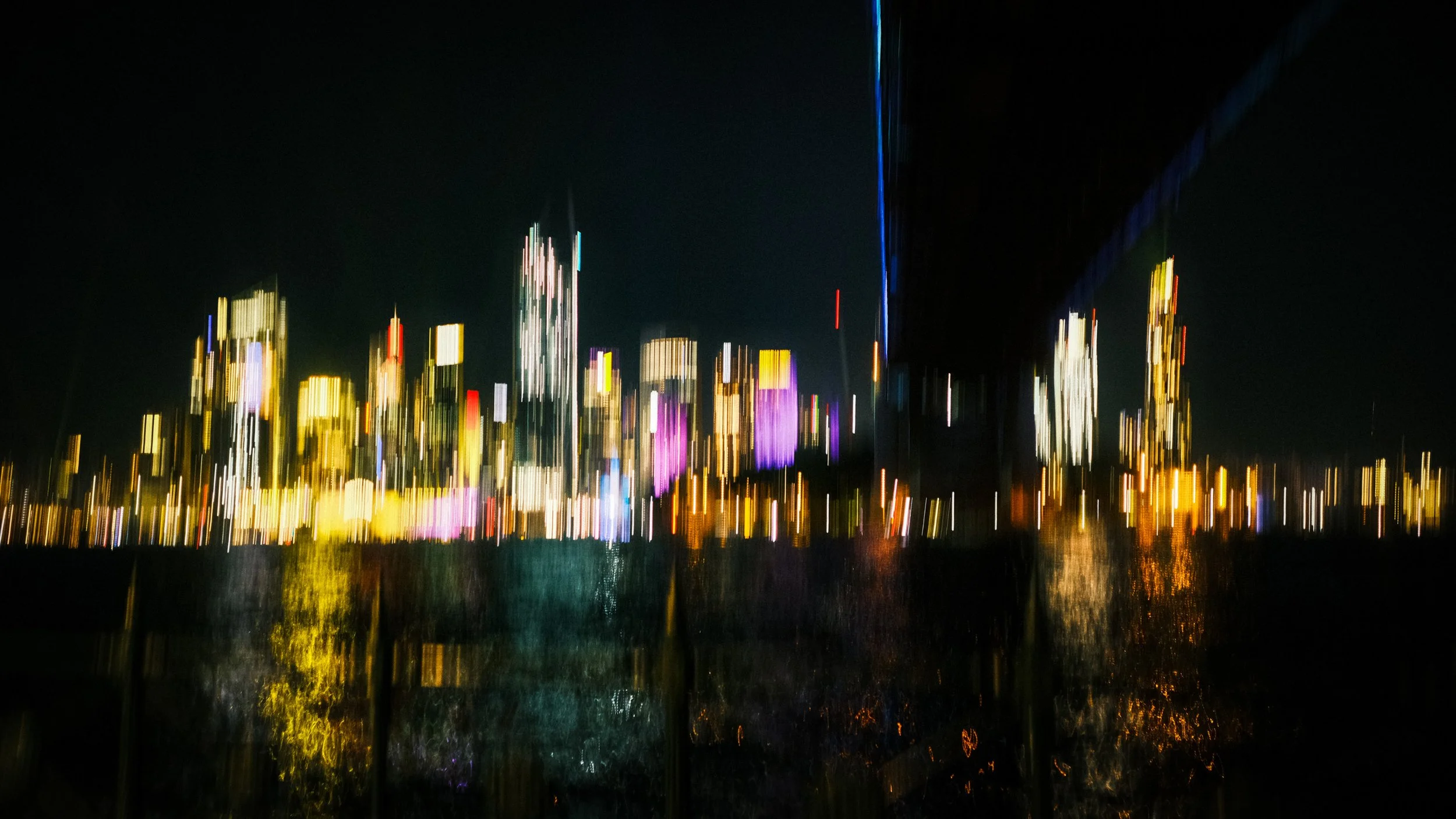 Blurred city skyline at night with colorful lights reflecting on water.