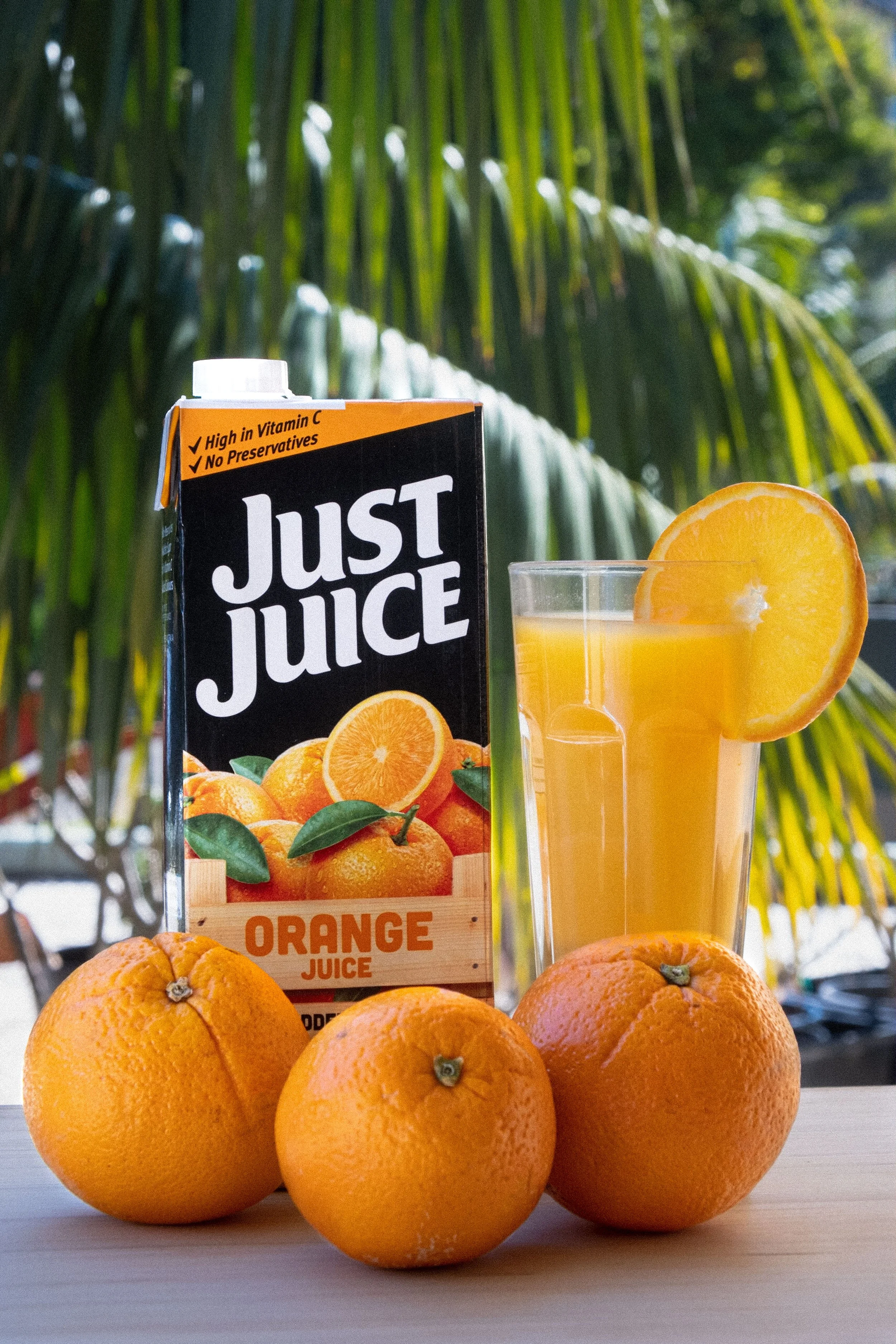 A carton of orange juice, a glass of orange juice with a slice of orange on the rim, and three whole oranges on a wooden surface, with a backdrop of green palm leaves.