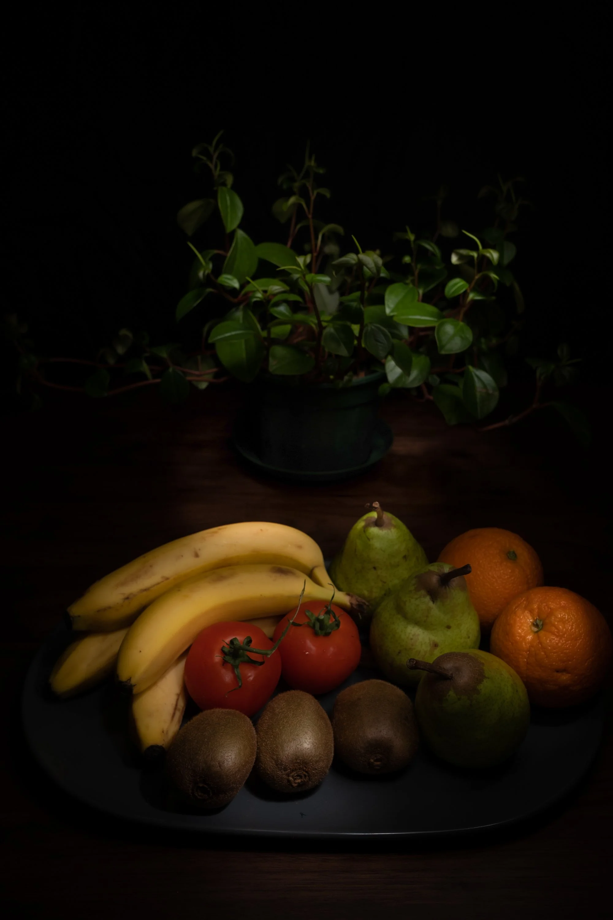 A black plate with bananas, tomatoes, kiwis, pears, and oranges, with a potted green plant in the background.