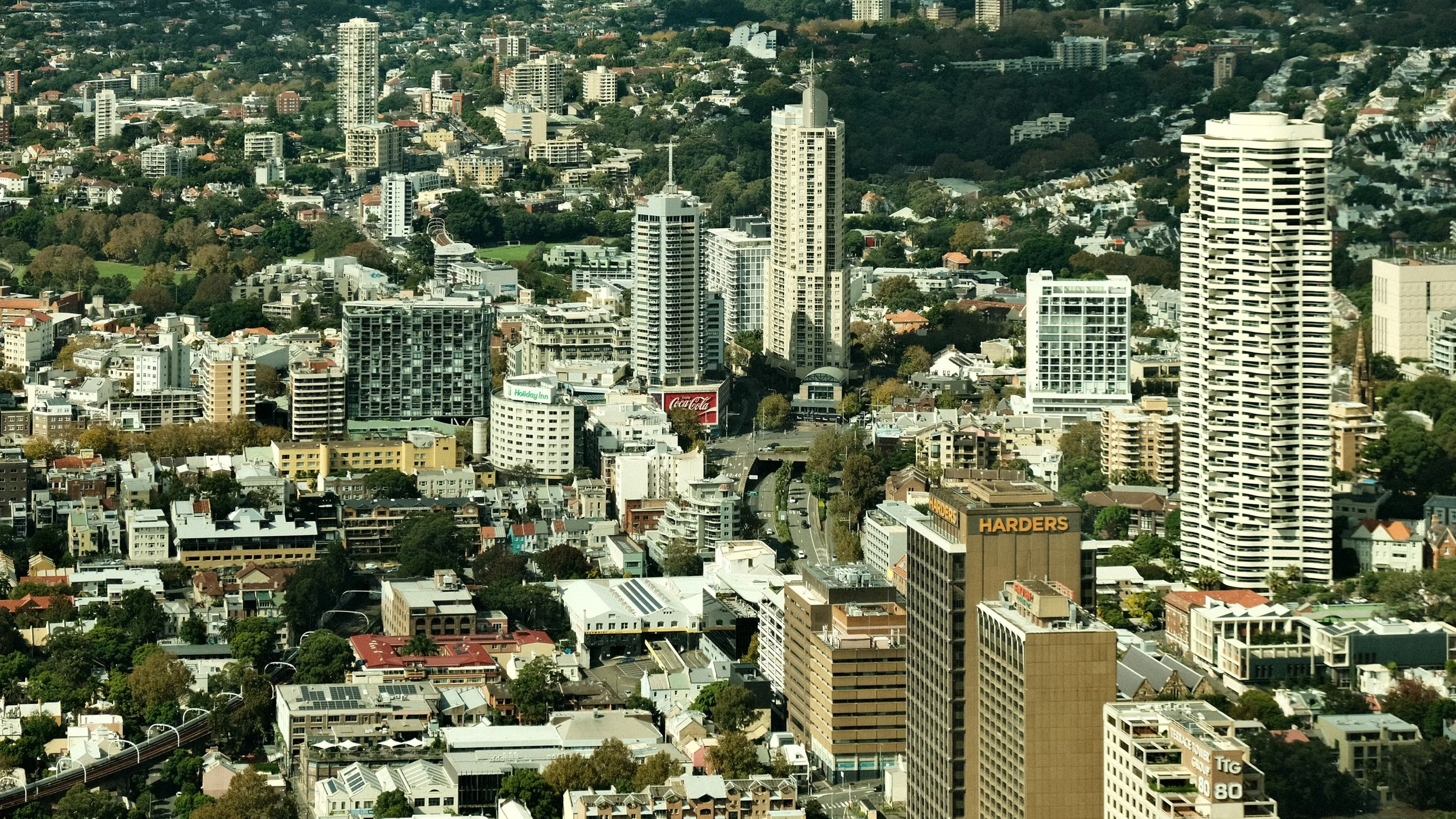 Aerial view of a city with a mix of high-rise buildings, mid-rise structures, and residential houses surrounded by trees, with a hilly landscape in the background.