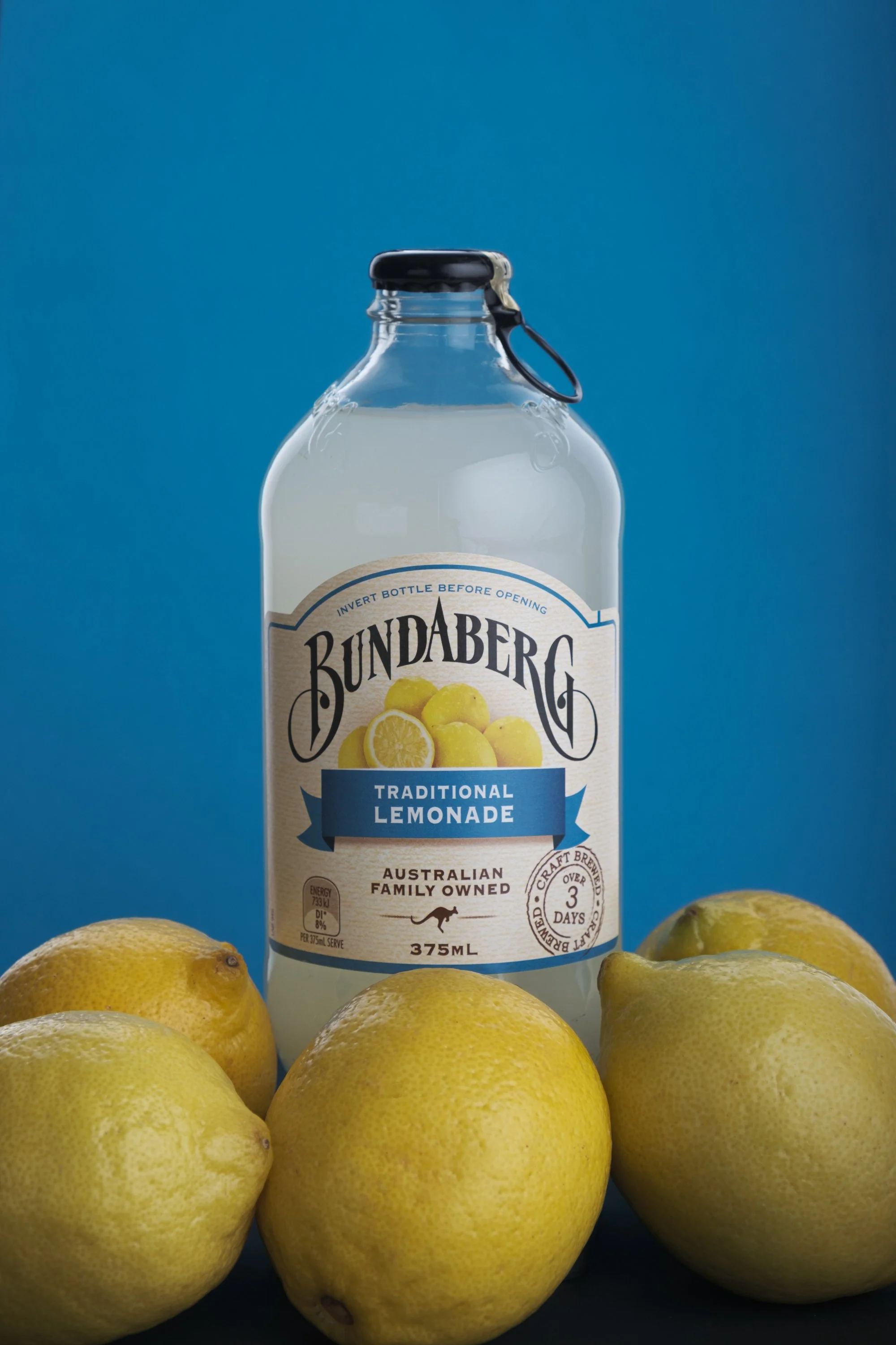 A bottle of Bundaberg traditional lemonade surrounded by fresh lemons on a dark surface with a blue background.