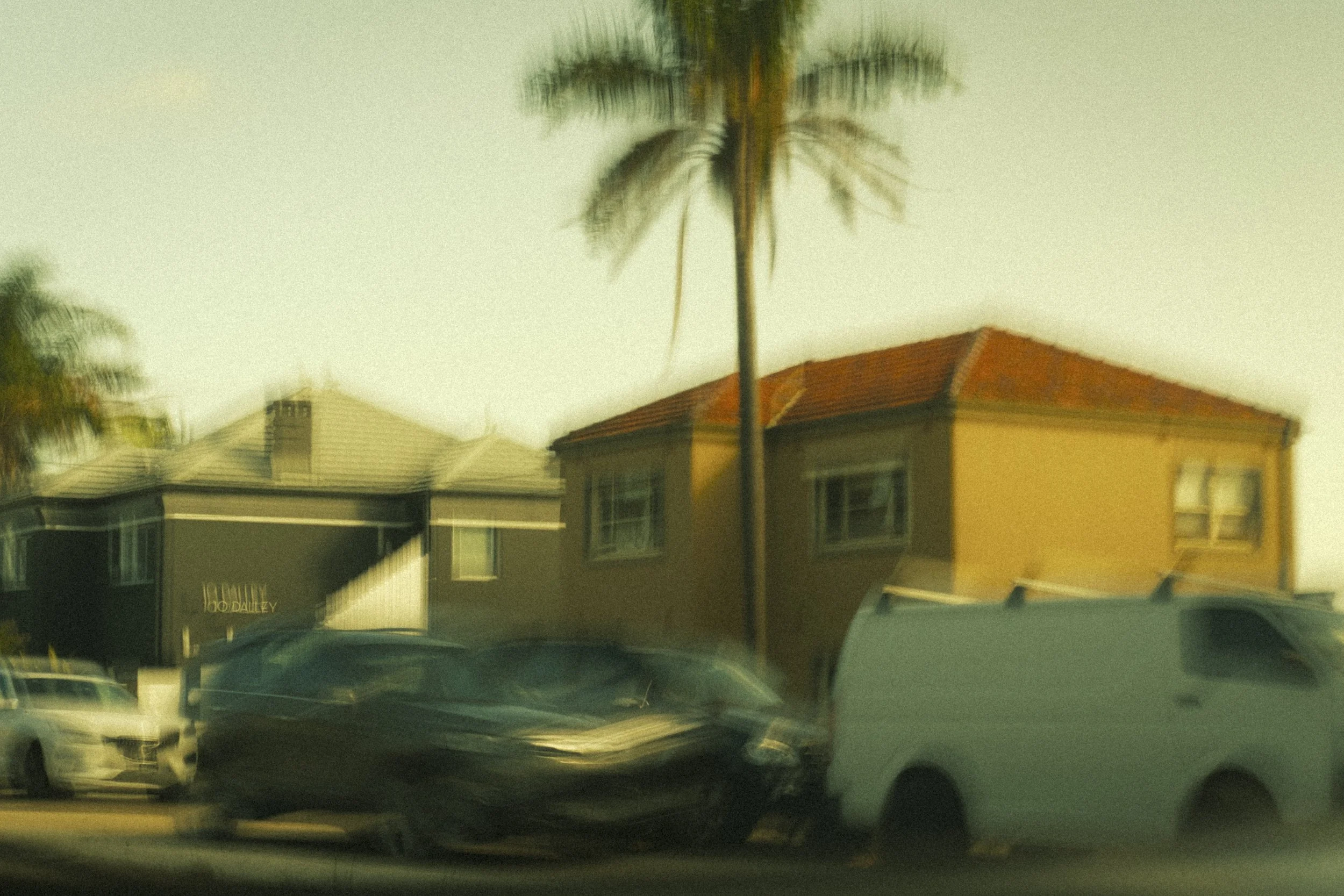 Blurred image of a street scene with parked cars, a tall palm tree, and residential buildings with red-tile roofs.