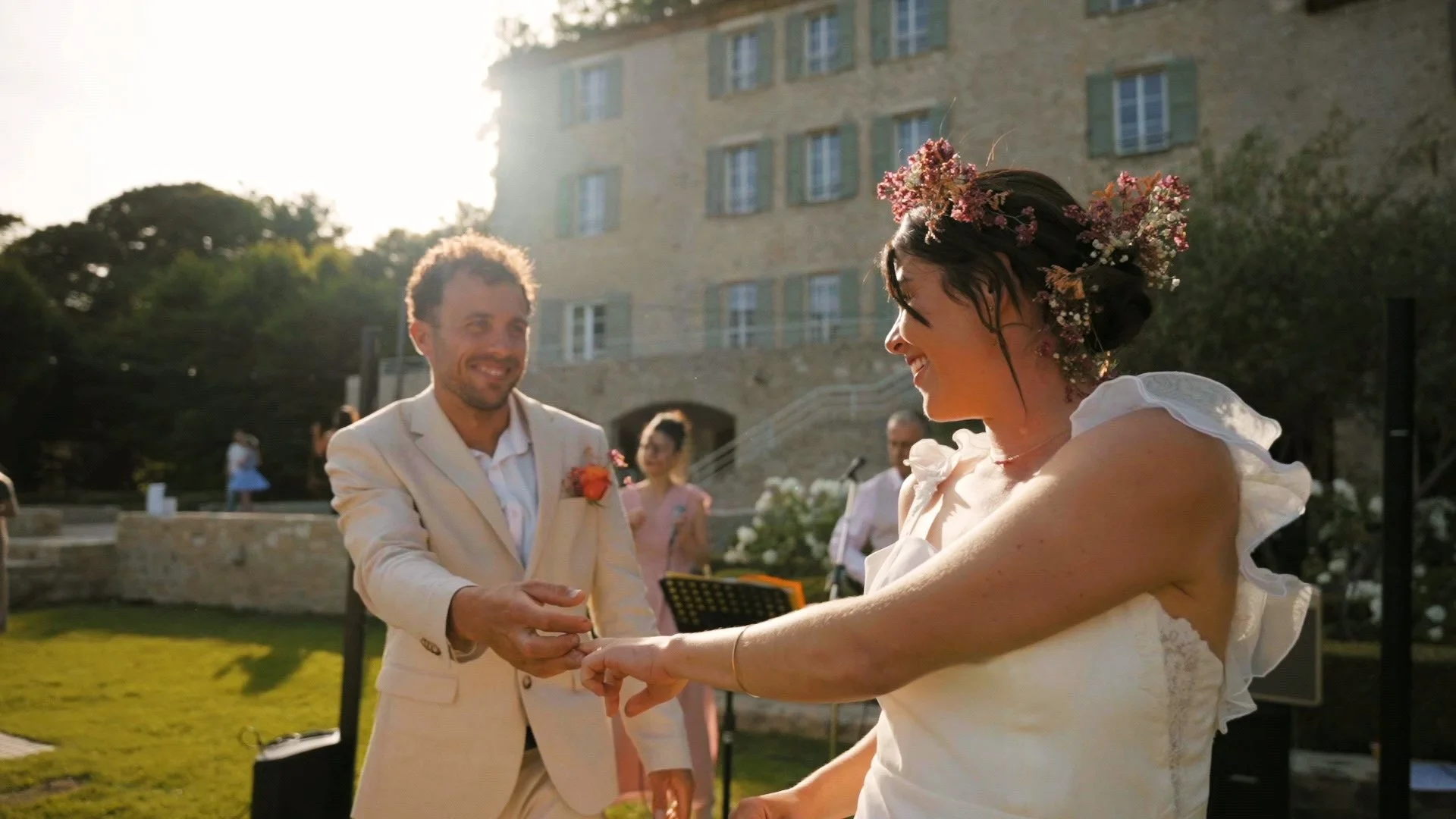 Un couple lors de leur mariage, la femme en robe blanche avec des fleurs dans les cheveux, l'homme en costume beige, en train d'échanger des alliances dans un jardin avec un bâtiment en pierre en arrière-plan, lors d'une cérémonie en plein air.