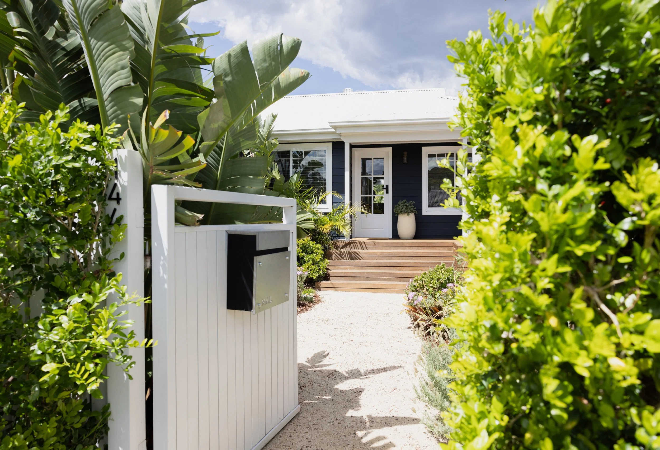 A beautifully presented coastal-style home framed by lush tropical landscaping and manicured hedges. A light-coloured pathway leads through the garden to a welcoming timber staircase and covered front porch.