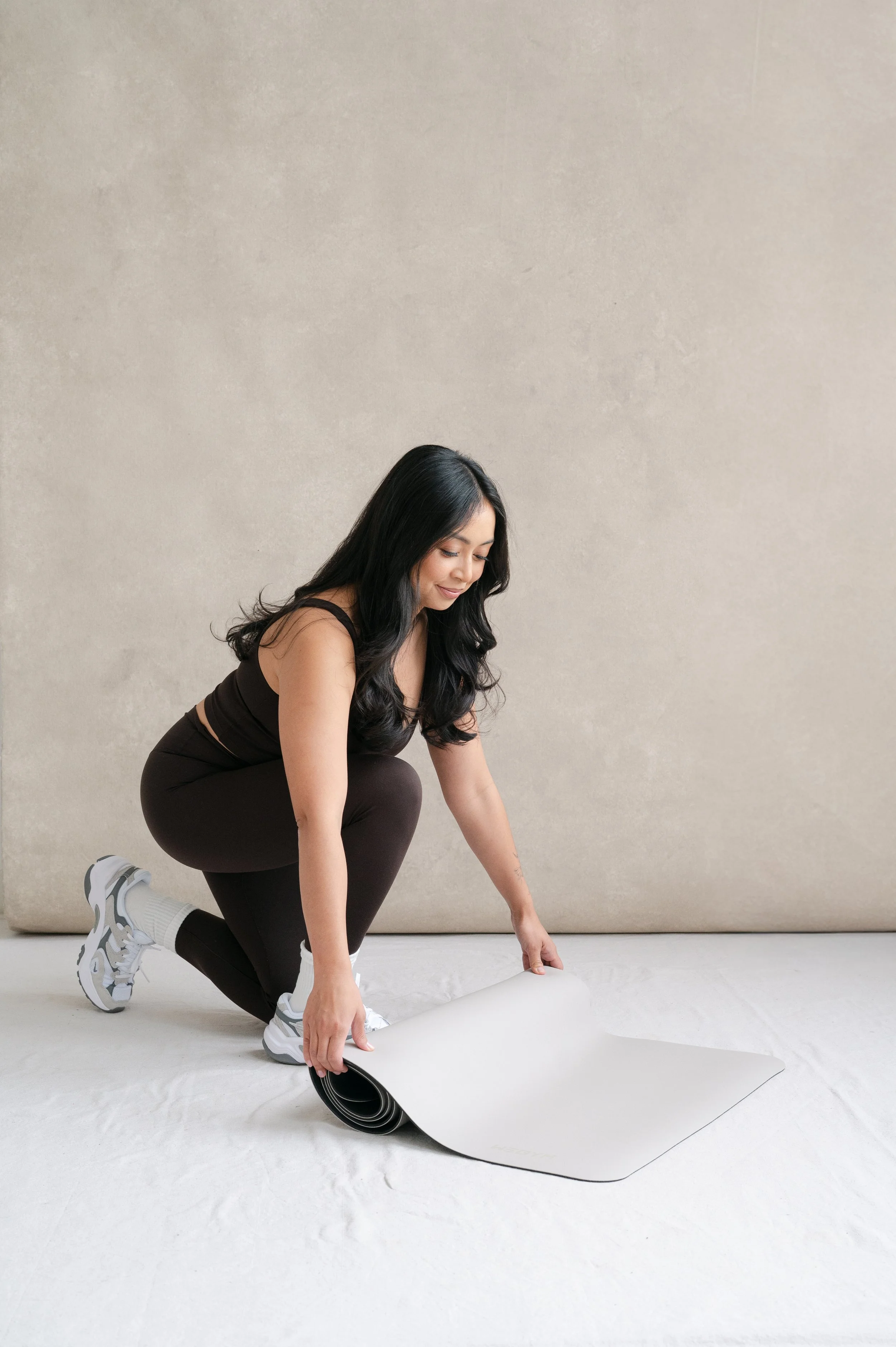 A woman with long black hair kneeling on the floor, unrolling a large white yoga mat.