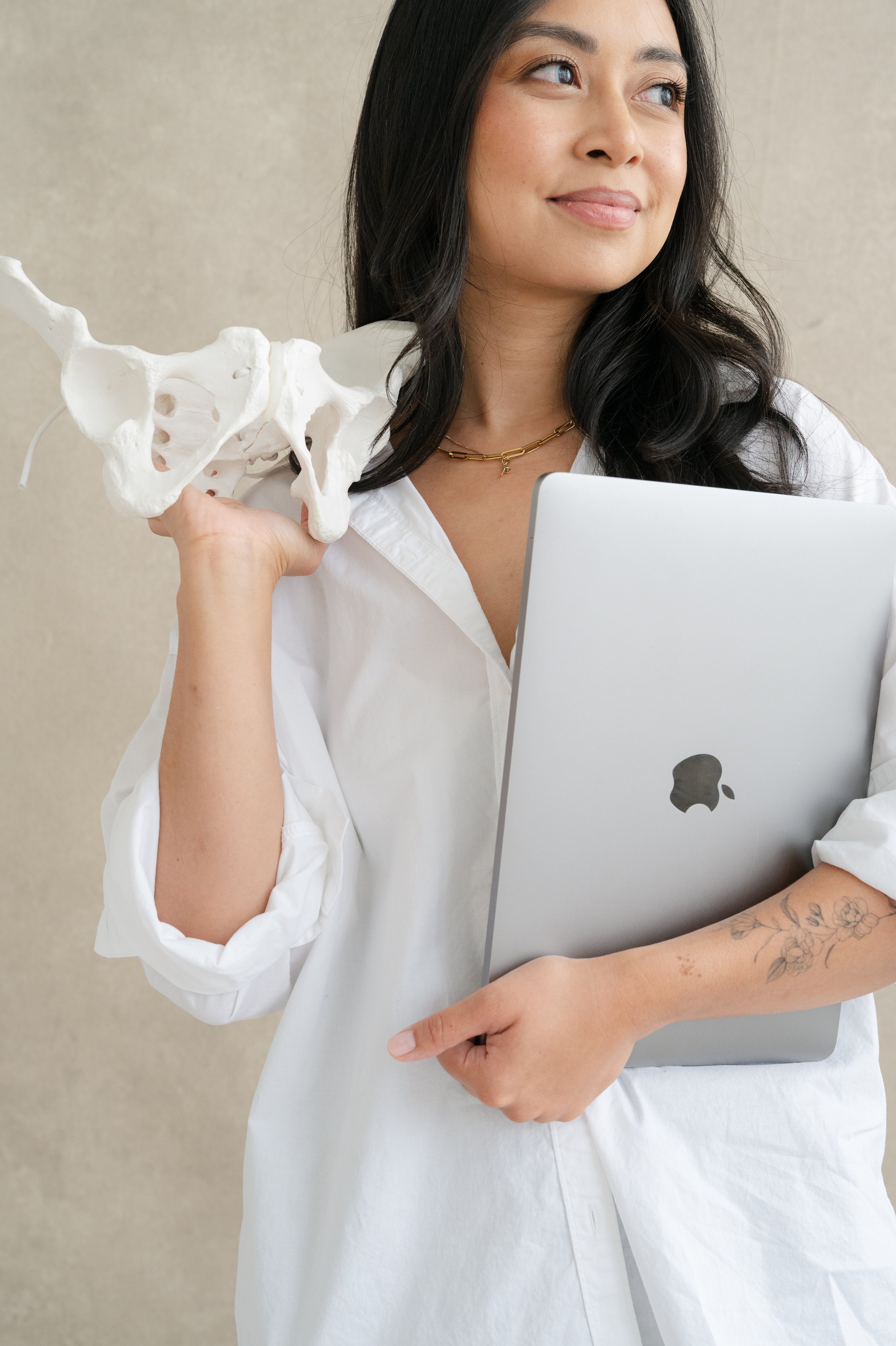 A woman with dark hair holding an anatomical skeleton pelvis model in her left hand and an open silver MacBook laptop under her right arm, standing against a beige background.